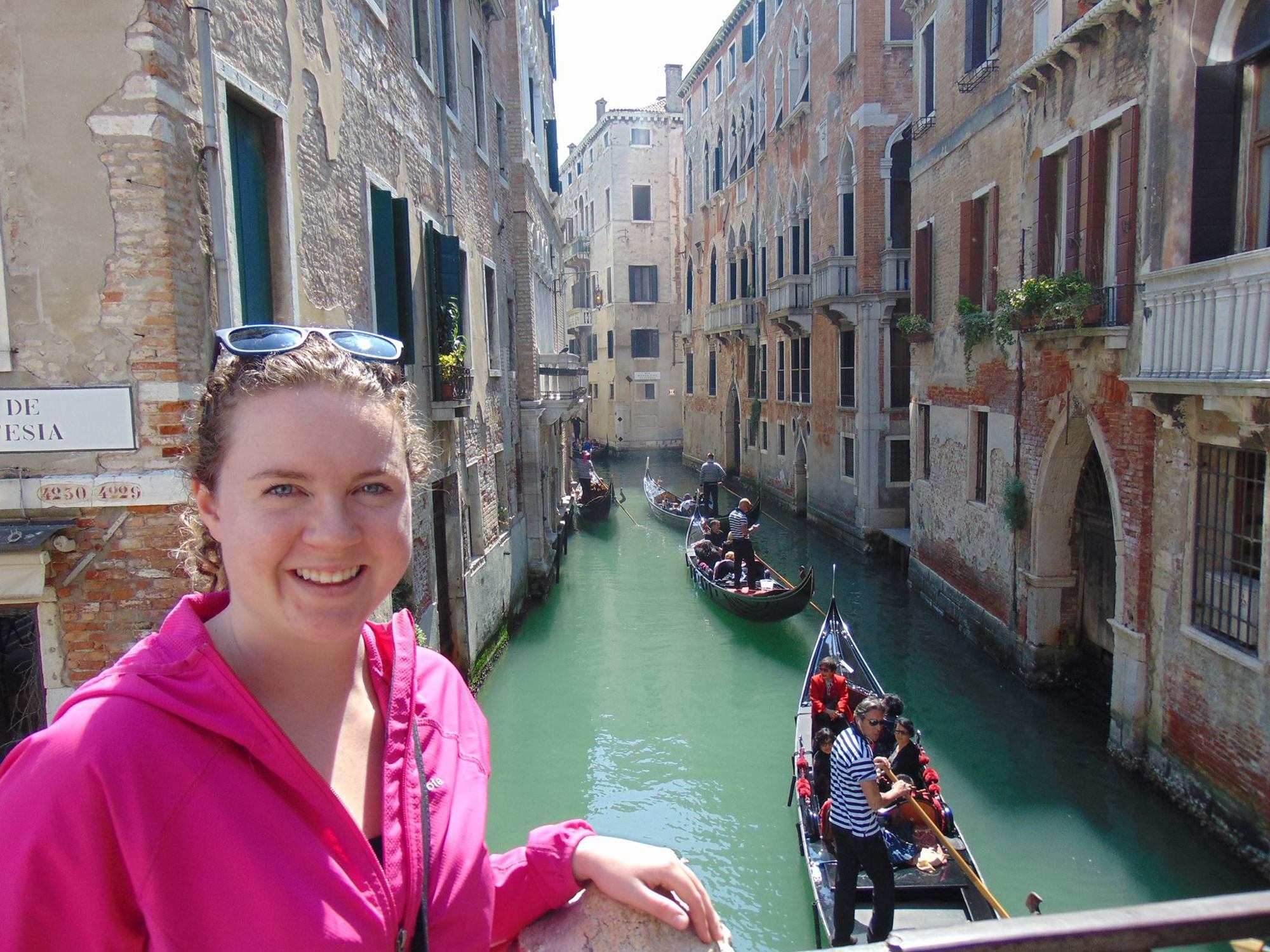 Autumn Fiske smiling at a Venice canal lined with historic brick buildings and gondolas passing below
