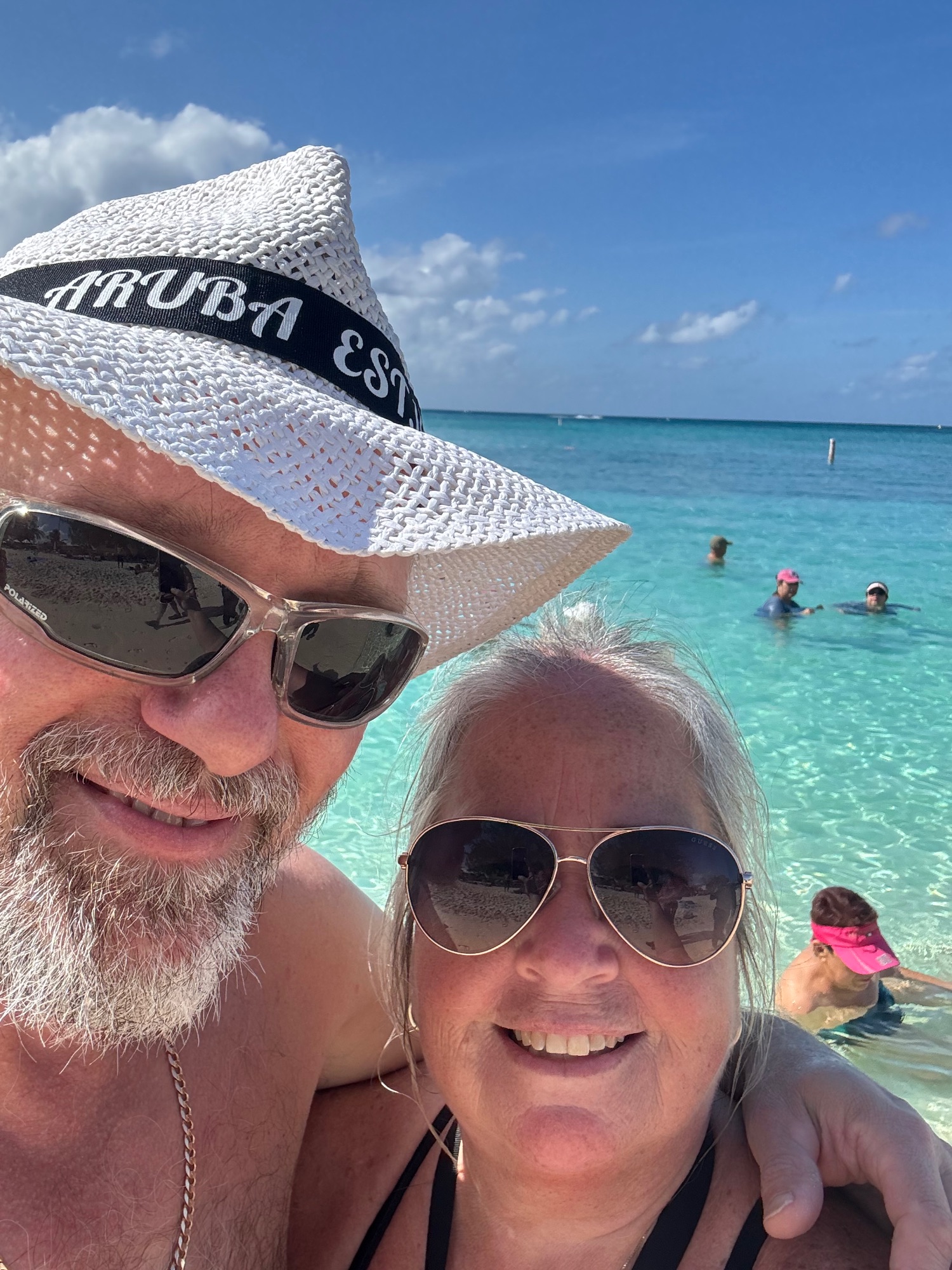 Couple in sunglasses and white hat smile together in turquoise Caribbean ocean water with swimmers in background