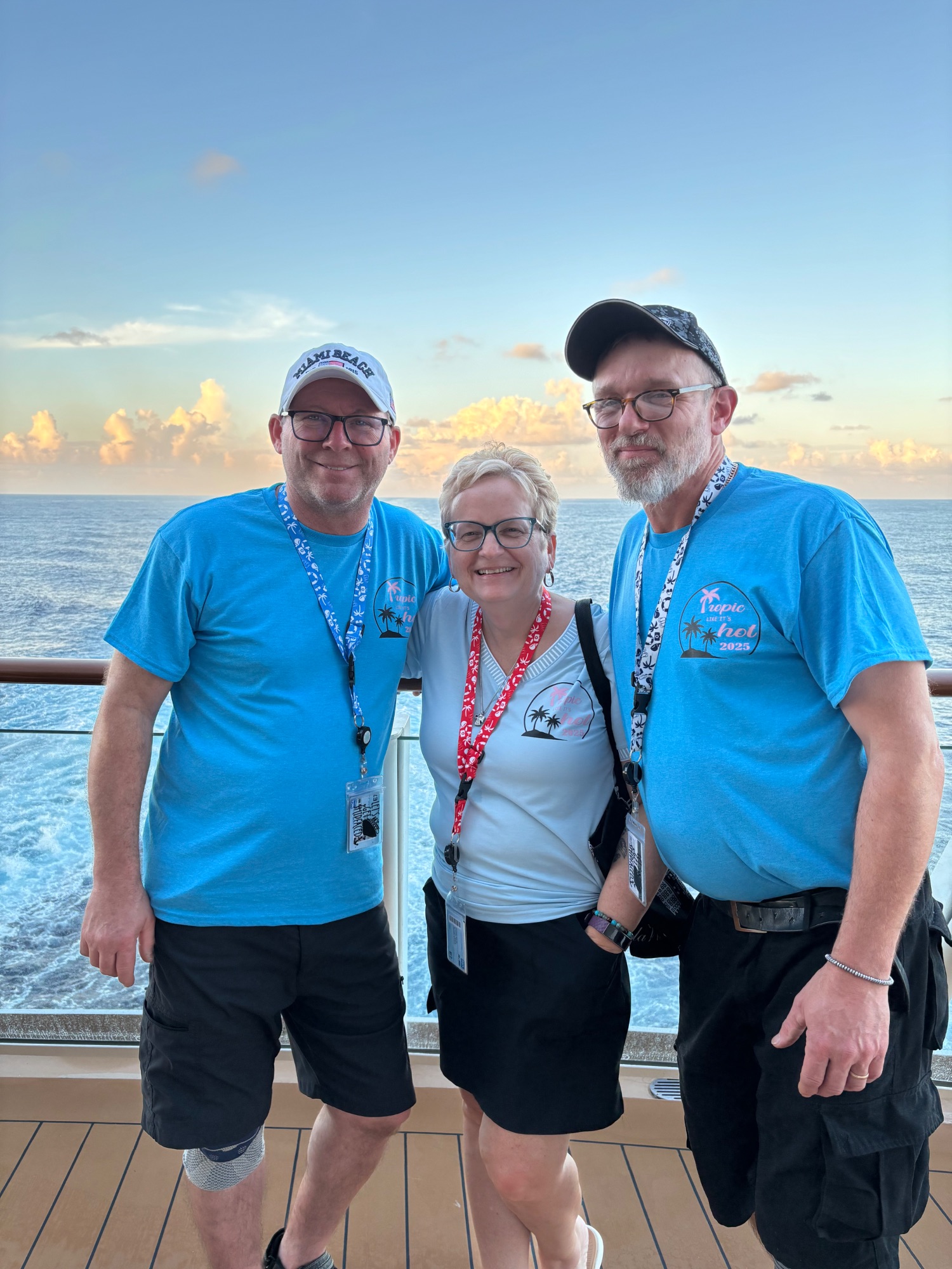 Three people wearing blue cruise shirts and lanyards smile on ship deck with ocean horizon at sunset