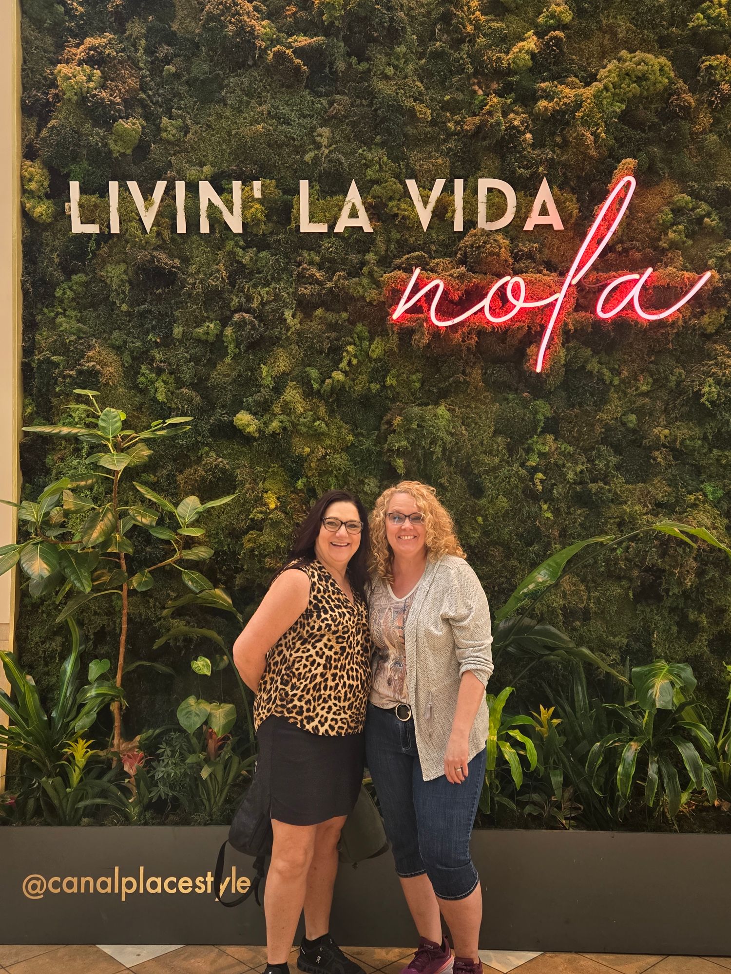 Two women pose together in front of a living moss wall with neon 'nola' sign at Canal Place in New Orleans