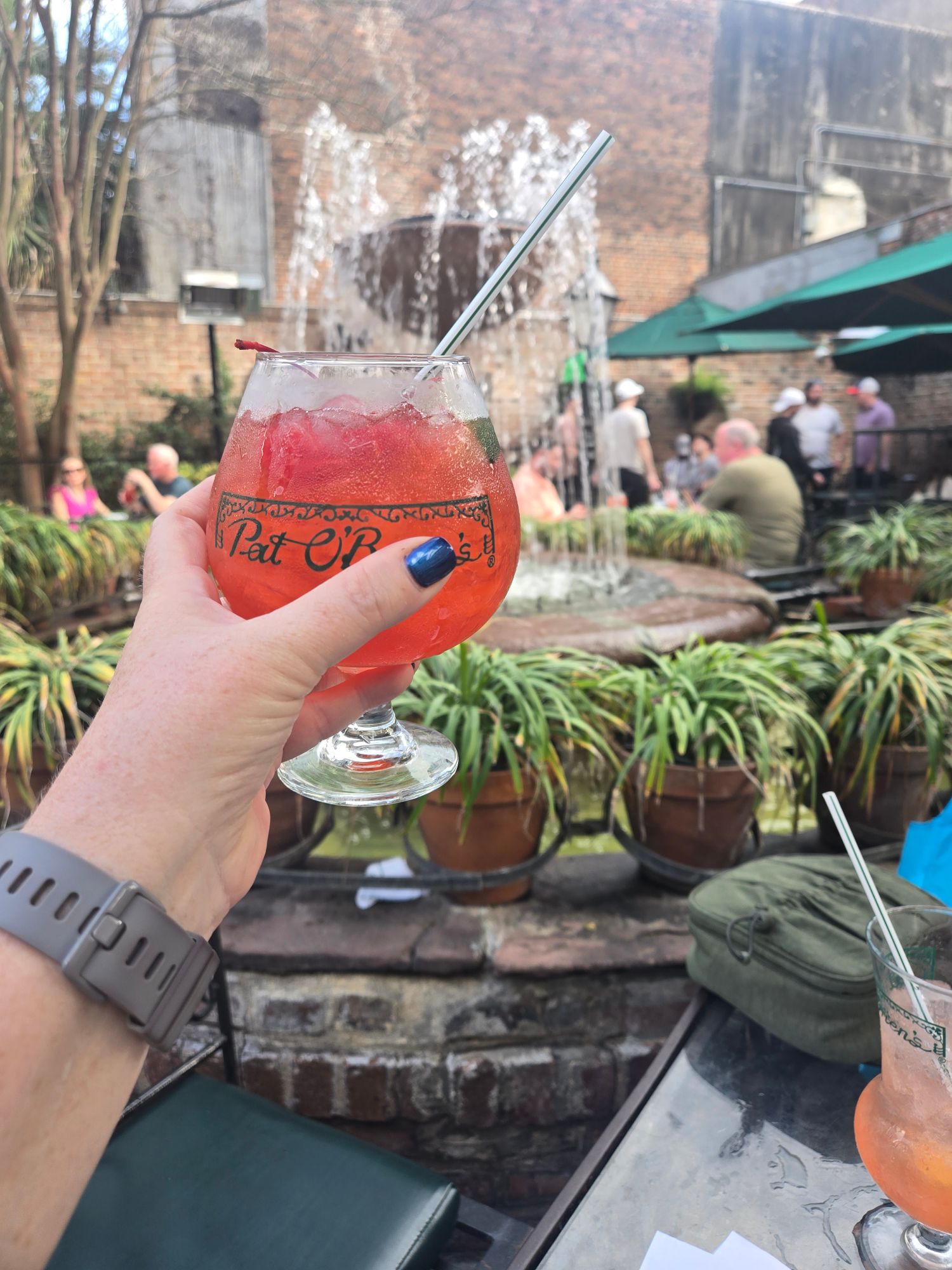 A hand holds a vibrant red cocktail with ice and a straw at an outdoor New Orleans courtyard venue with brick buildings