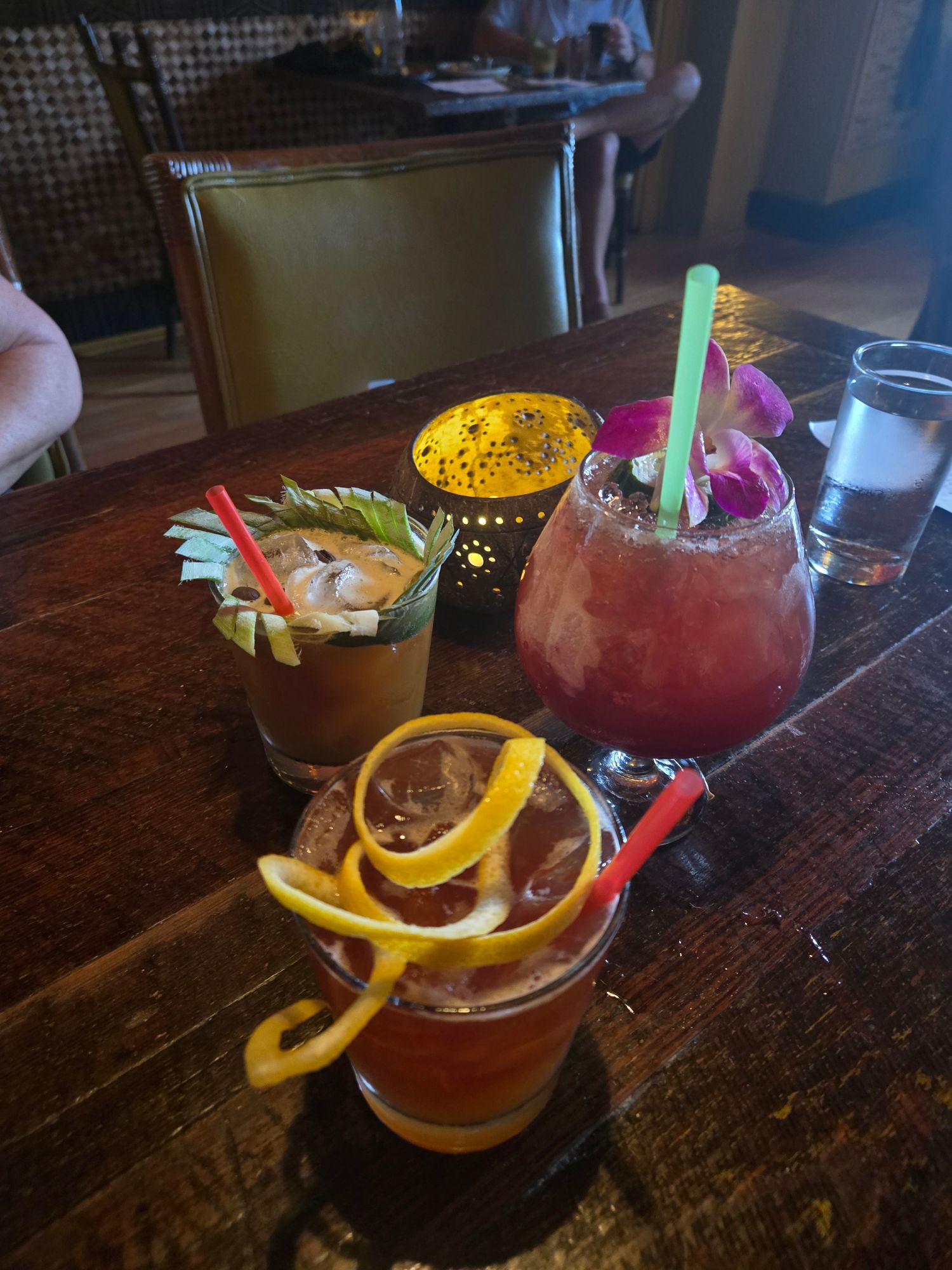 Three colorful tropical cocktails on a dark wooden bar counter, garnished with citrus, pineapple, and orchid flowers