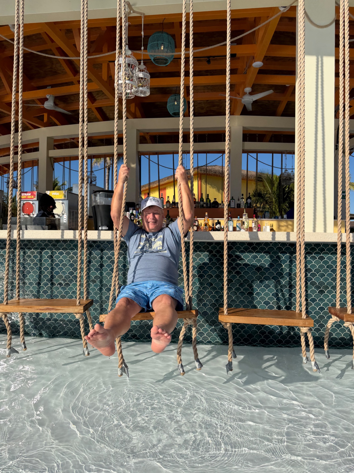 Man on rope swing in shallow turquoise water with ship visible in background