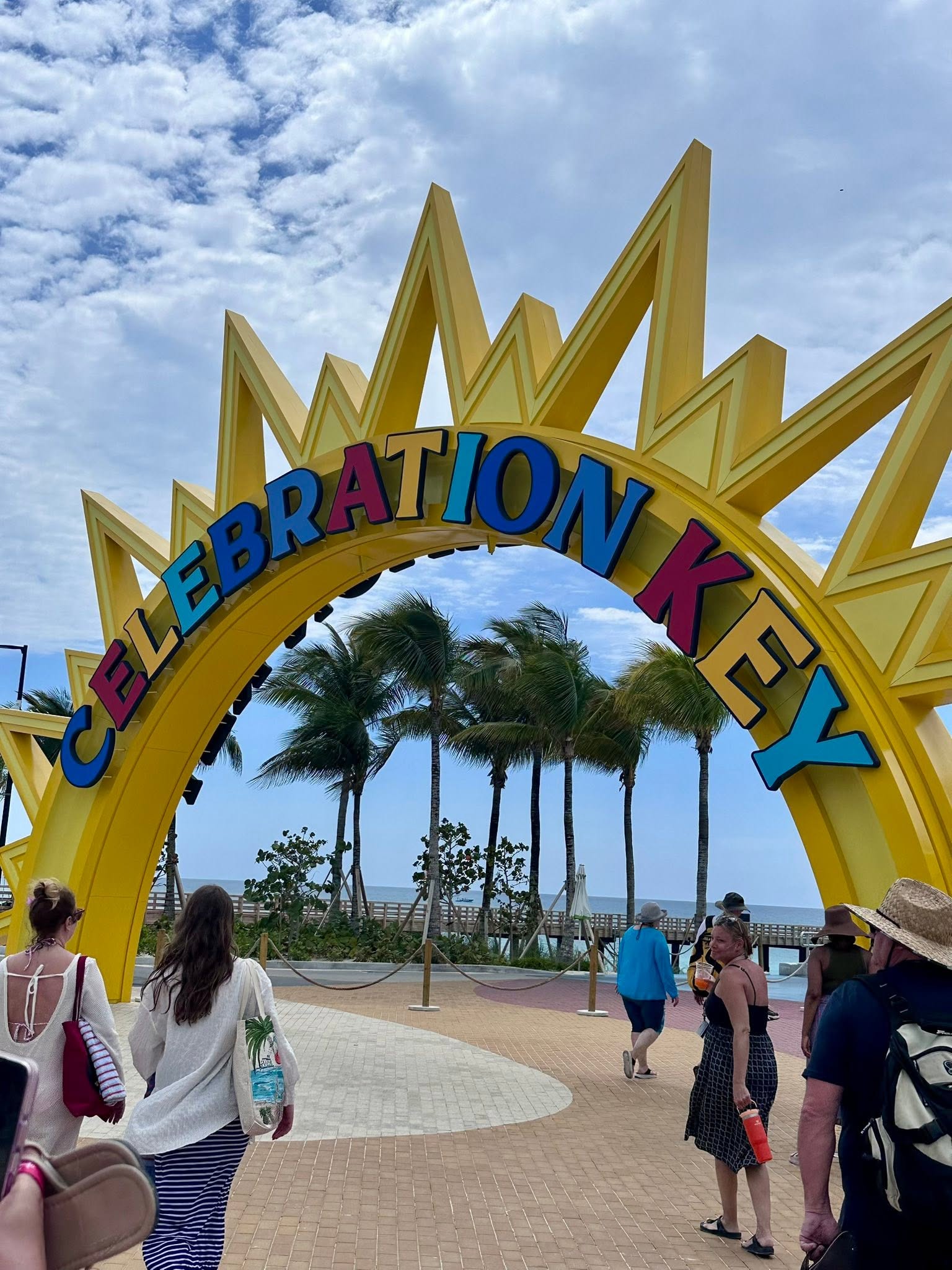 Large yellow Celebration Key arch with colorful letters and palm trees, visitors walking on blue plaza near beach