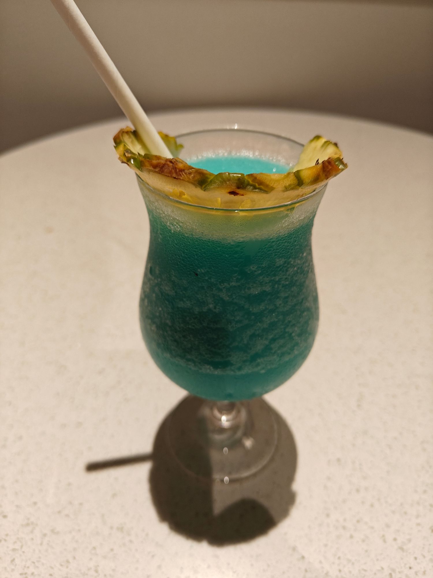 Turquoise cocktail with charred pineapple garnish and white straw in a glass on white surface