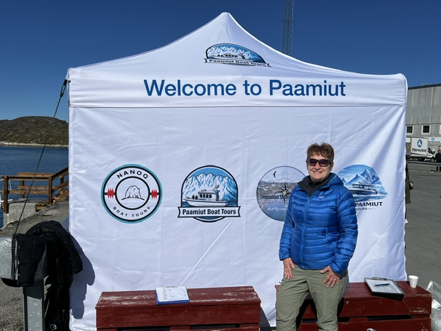 Woman in blue jacket stands in front of a white tent banner reading 'Welcome to Paamiut' at a harbor