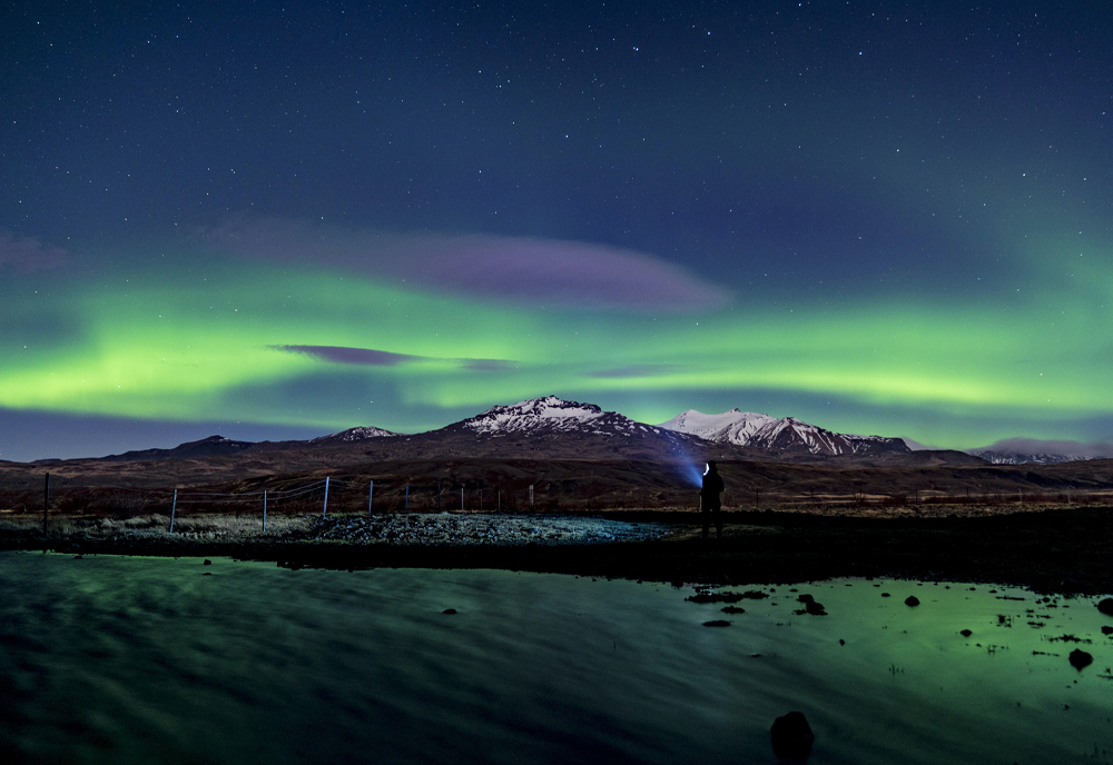 Northern lights shimmer green above snow-capped mountains in Iceland, with a silhouetted figure standing by a river in the foreground