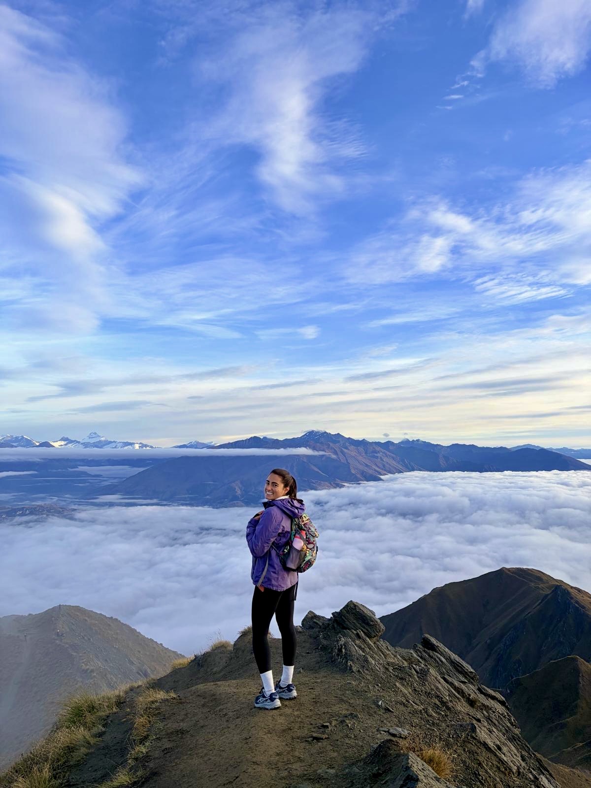 Julia standing on a mountain ridge at sunrise with cloud layers below and distant peaks visible across the landscape