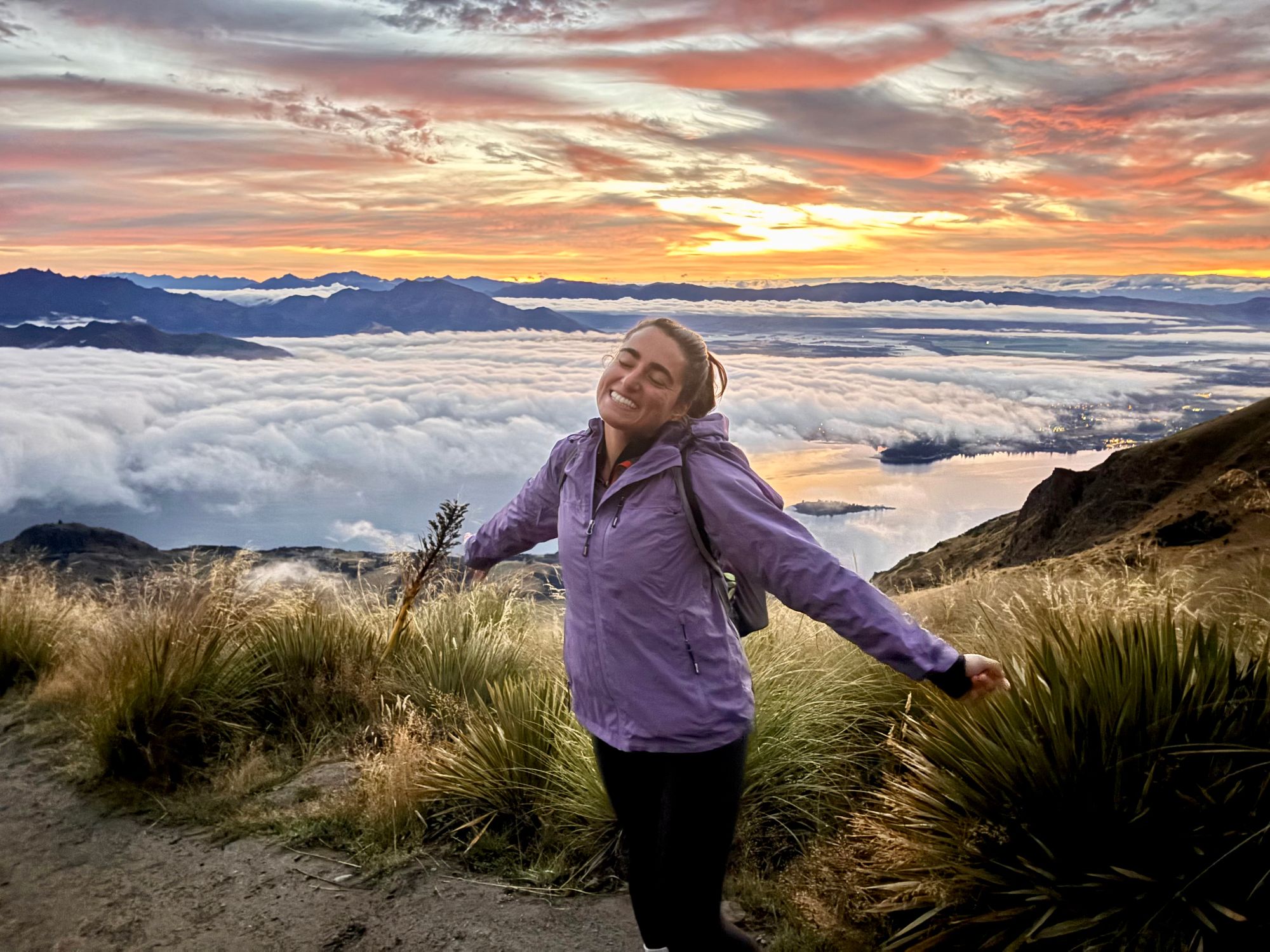 Julia in a purple jacket celebrating at sunset on a grassy hilltop overlooking a cloud-covered valley and distant mountains