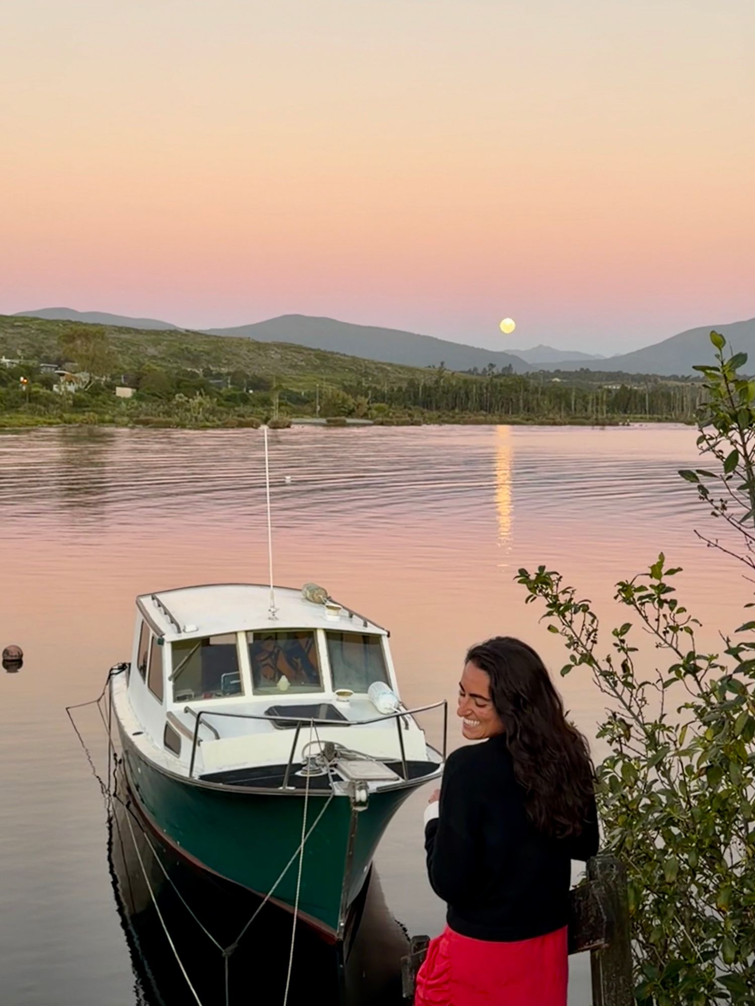 Julia smiling beside a moored fishing boat at dusk with mountains and a full moon reflected in calm water