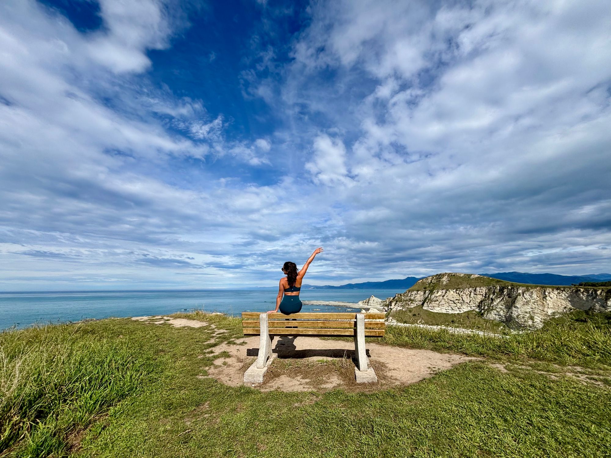 Julia sitting on a wooden bench on a coastal cliff edge with ocean, islands, and mountains visible in the distance