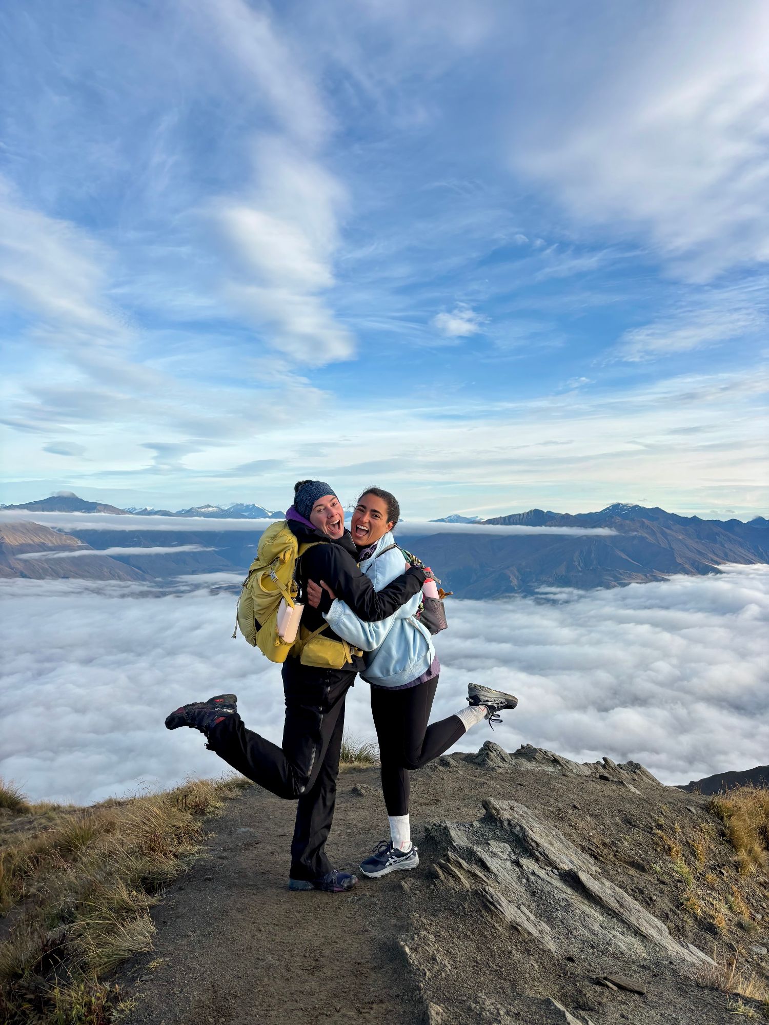 Julia and a friend hugging on a mountain summit above clouds with snow-capped peaks visible across the landscape