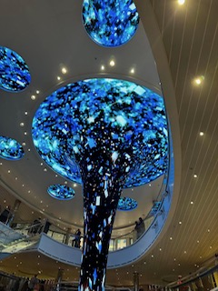 Illuminated blue tree sculpture with glowing canopy suspended in a modern indoor atrium space