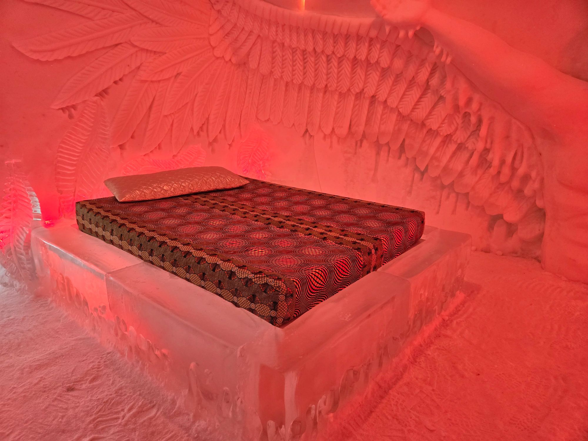 A decorative bed with patterned red bedding sits on an ice platform inside a room with red-tinted ice walls and carved details