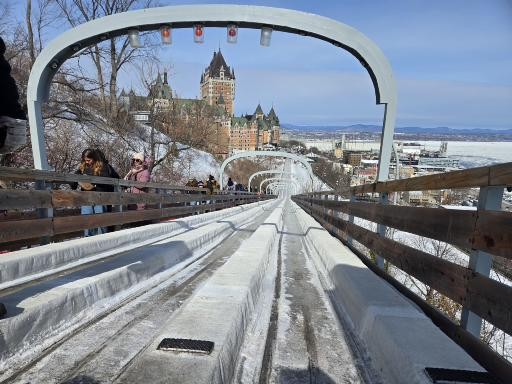 View down an icy bobsled track with wooden railings, showing Château Frontenac and Quebec City landscape in the distance