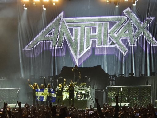 Anthrax performs on stage with purple and white lighting and their logo displayed on the backdrop at a concert venue