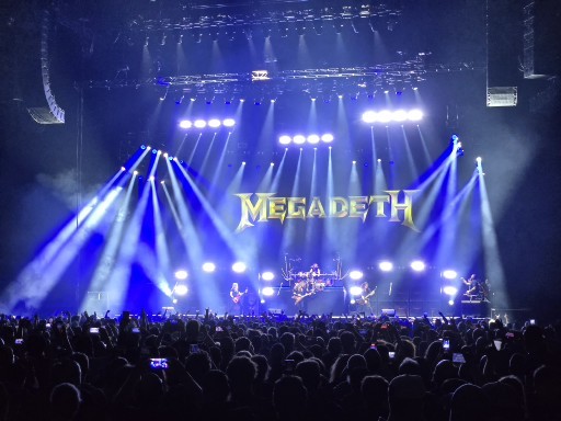 Megadeth performs on stage with blue stage lighting, their name displayed in gold letters, and a large crowd in the foreground