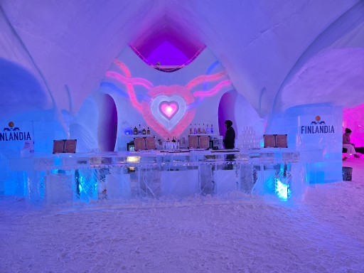 Interior of an ice hotel bar with purple and pink neon lighting, ice furniture, and a heart-shaped neon sign on the wall