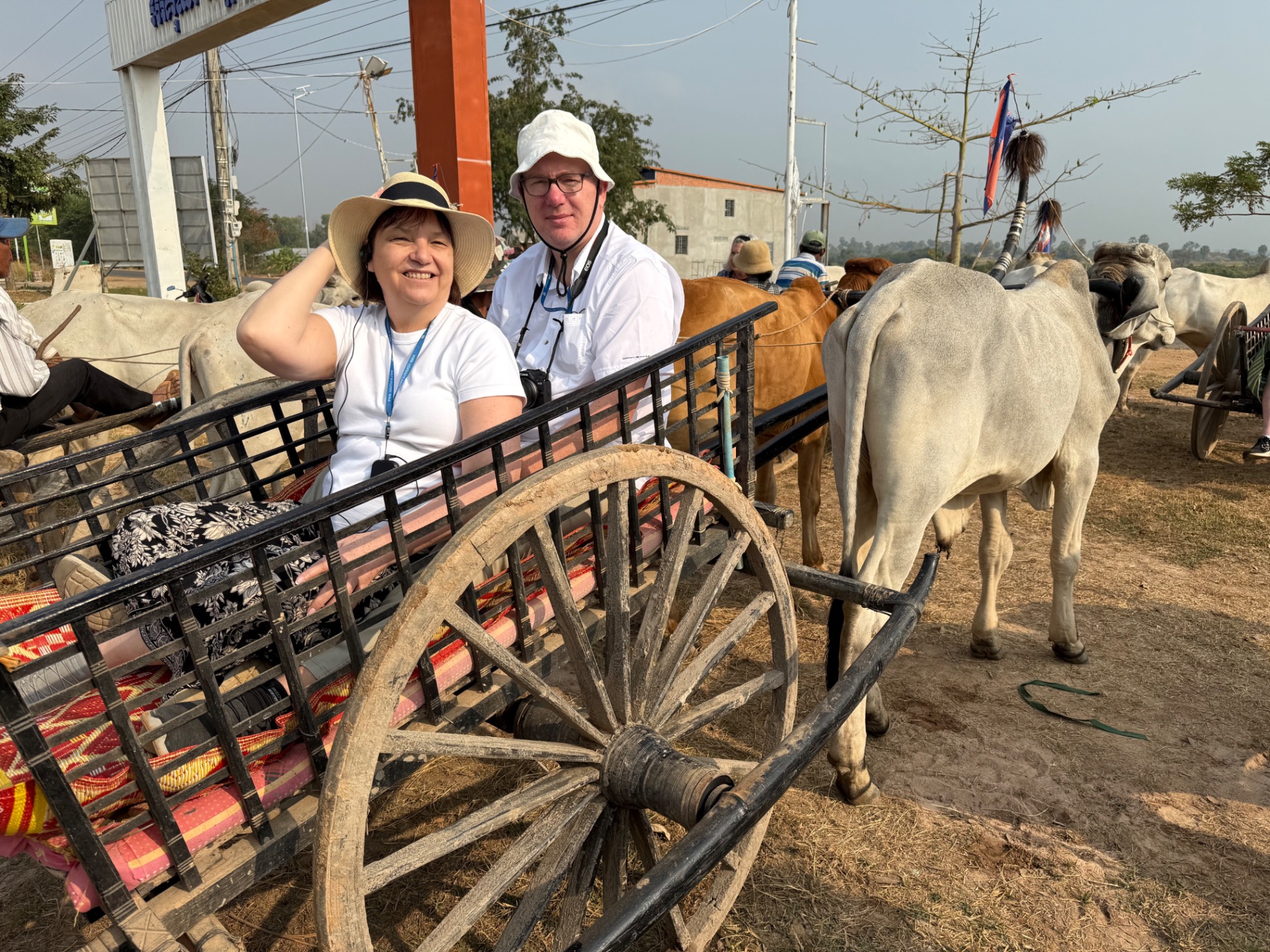 Two people smiling at camera beside a traditional wooden ox-drawn cart at a rural livestock market