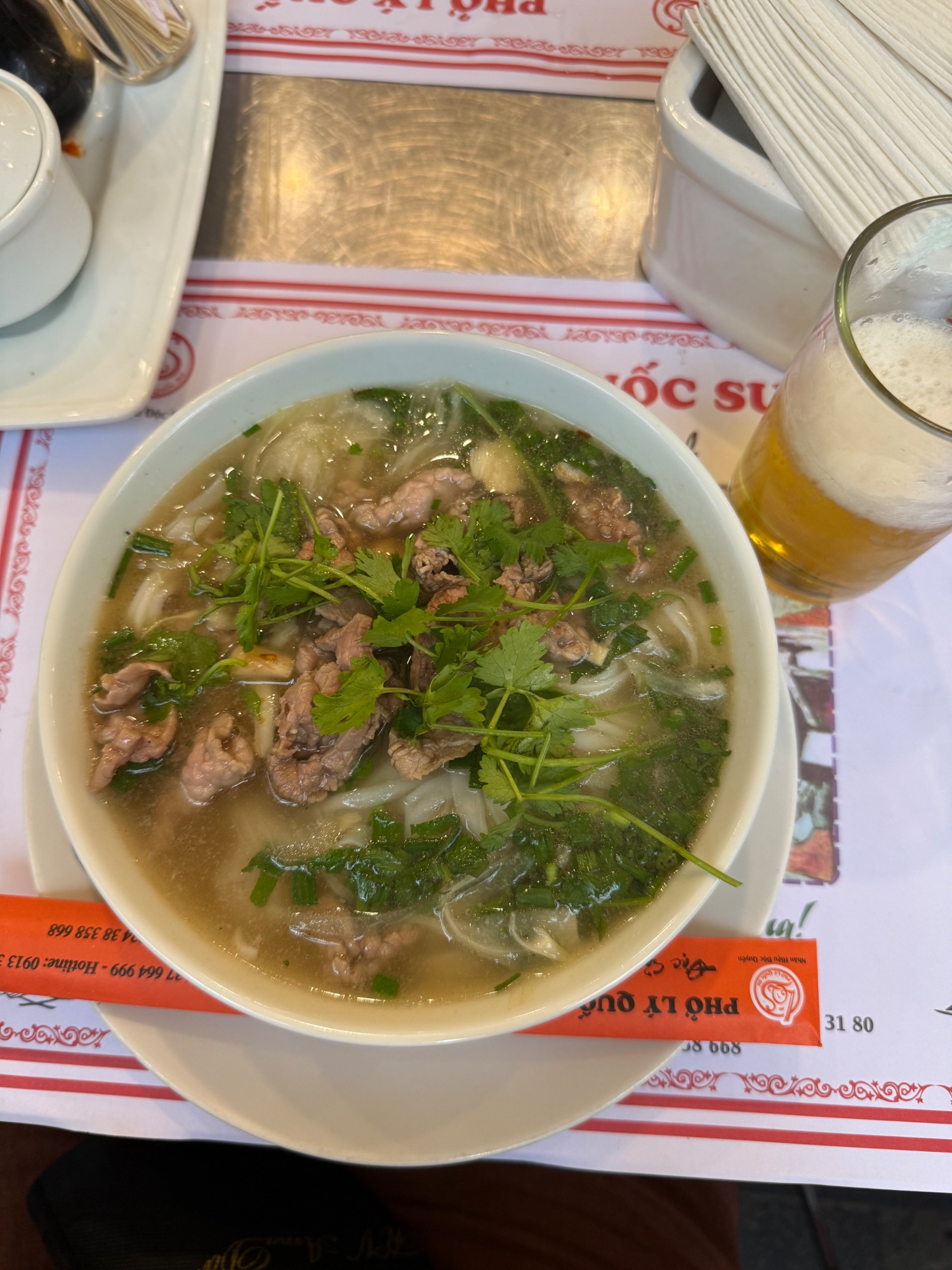 Bowl of Vietnamese pho with beef, noodles and fresh herbs alongside beer on restaurant table