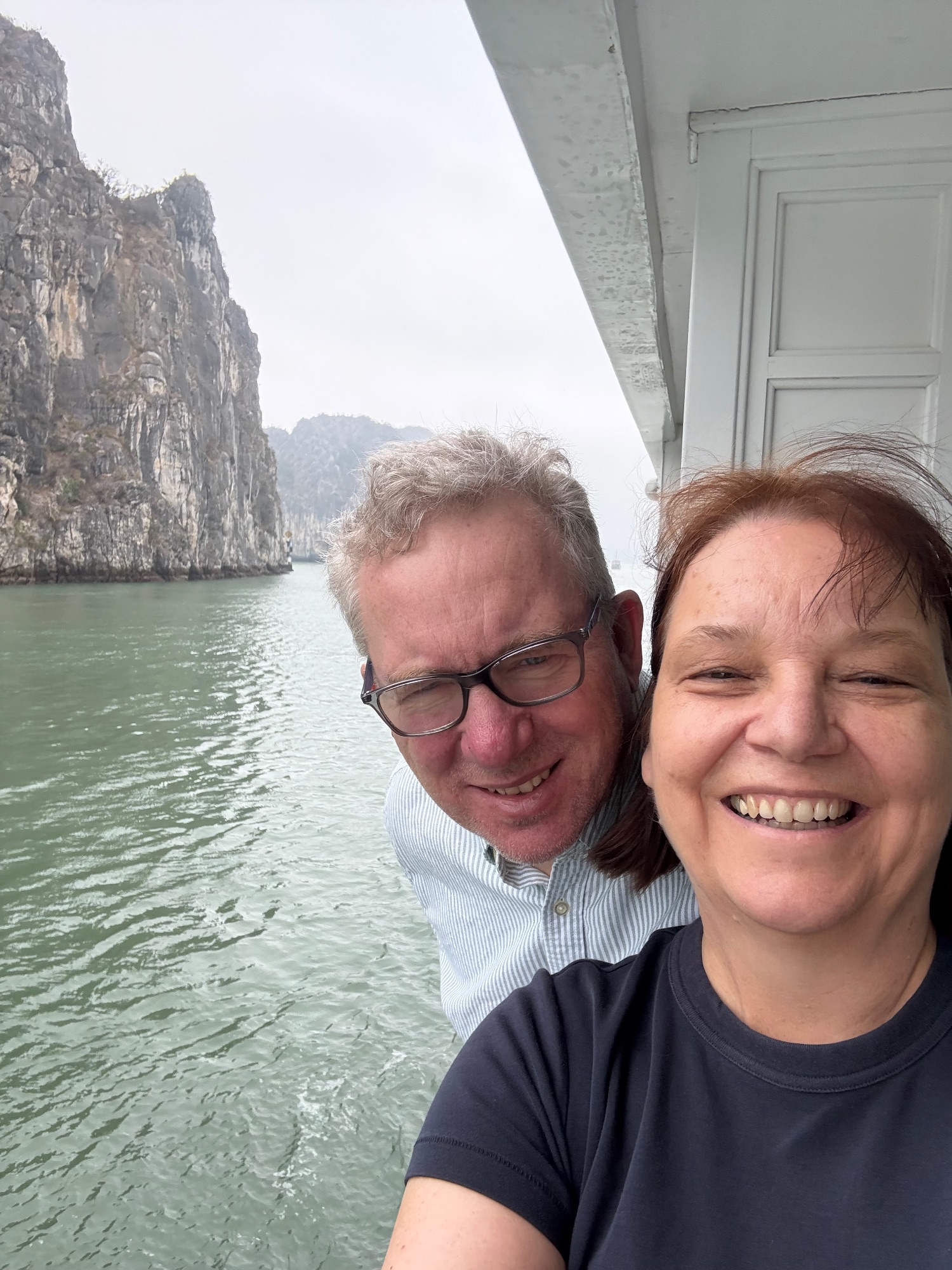 Two people smiling at camera on boat with dramatic limestone cliffs and junks in Halong Bay background