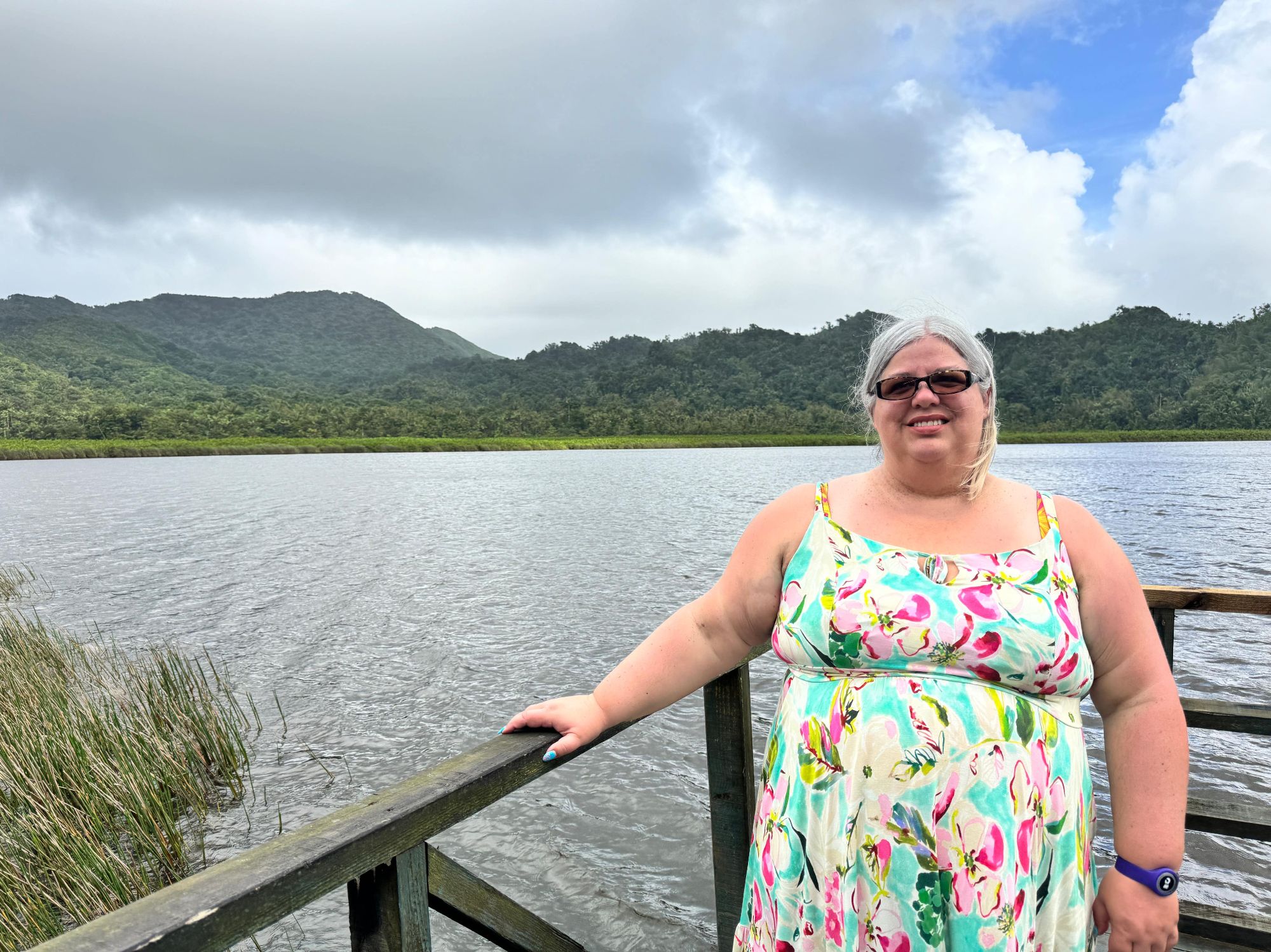 A smiling woman in a floral dress poses on a boardwalk overlooking a tropical lagoon backed by rainforest mountains.