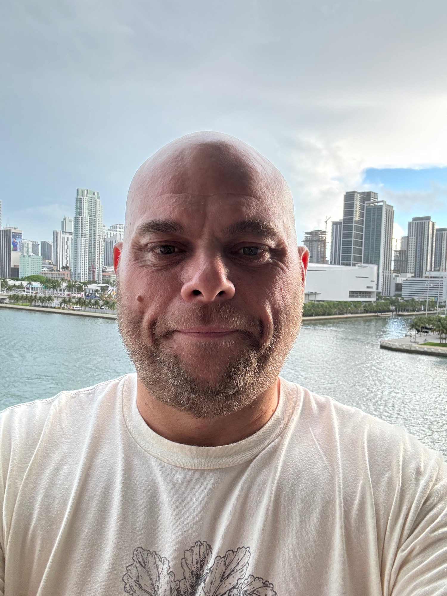 Man in beige t-shirt smiling at camera with a coastal city skyline and waterway behind him