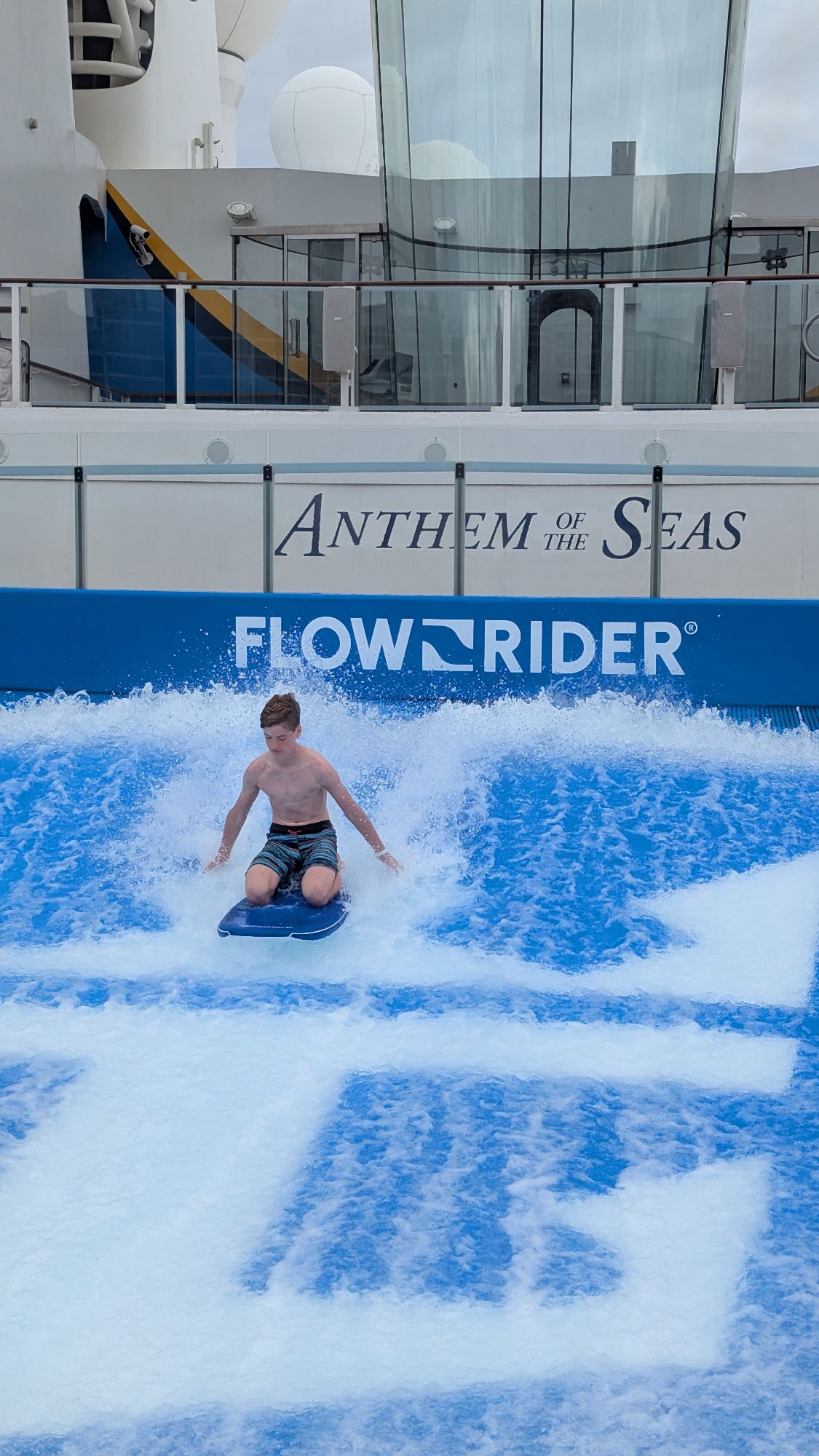 Young boy surfing on FlowRider wave simulator aboard Anthem of the Seas with ship's hull visible behind