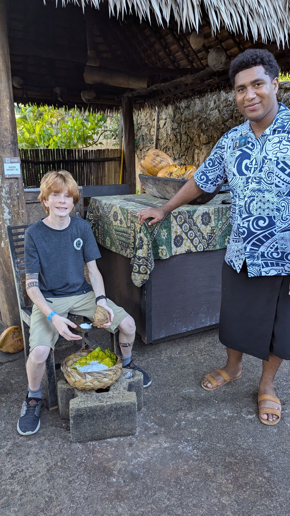 Young boy with local Fijian man in traditional patterned shirt at tiki bar with woven baskets and coconuts