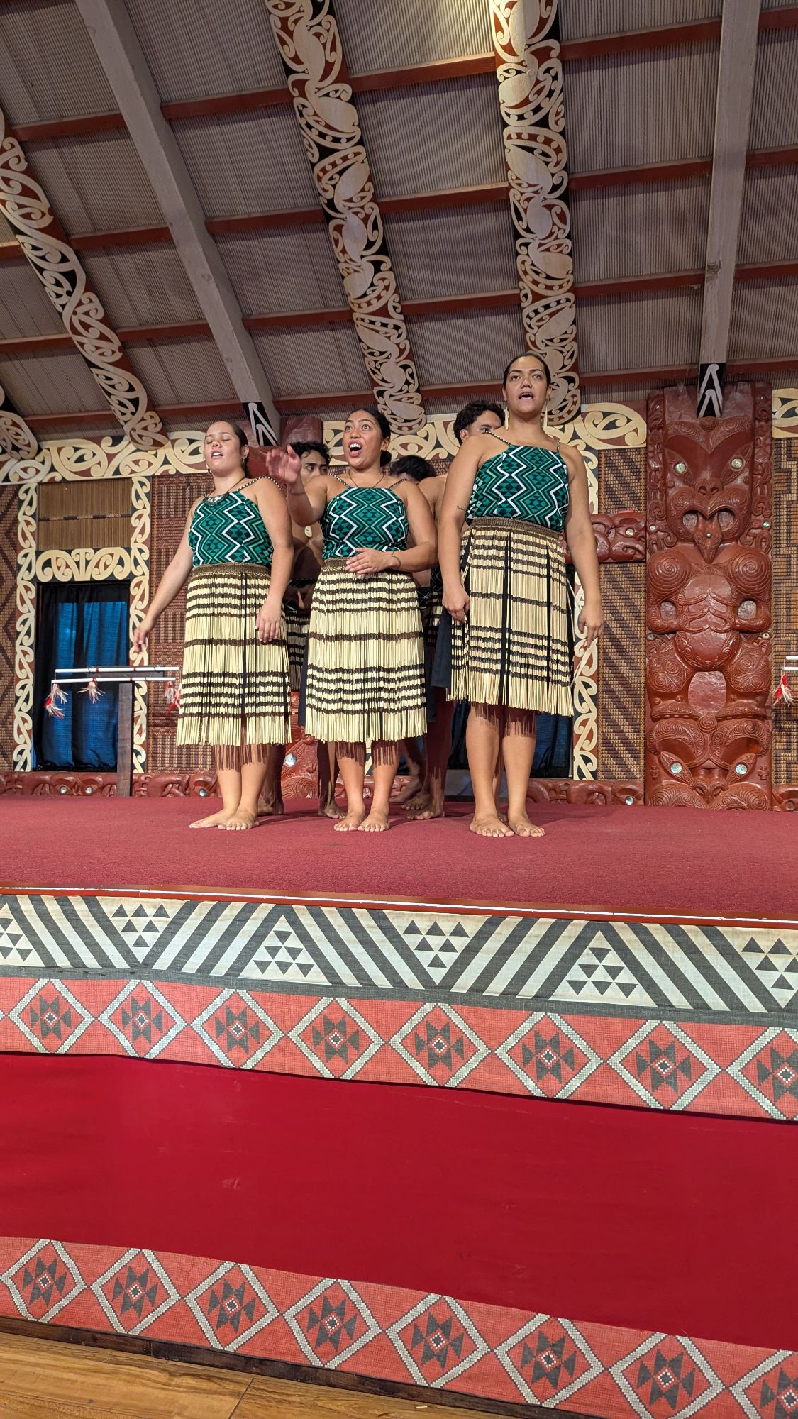 Maori women performing haka in traditional green and cream woven skirts on stage with carved wooden panels