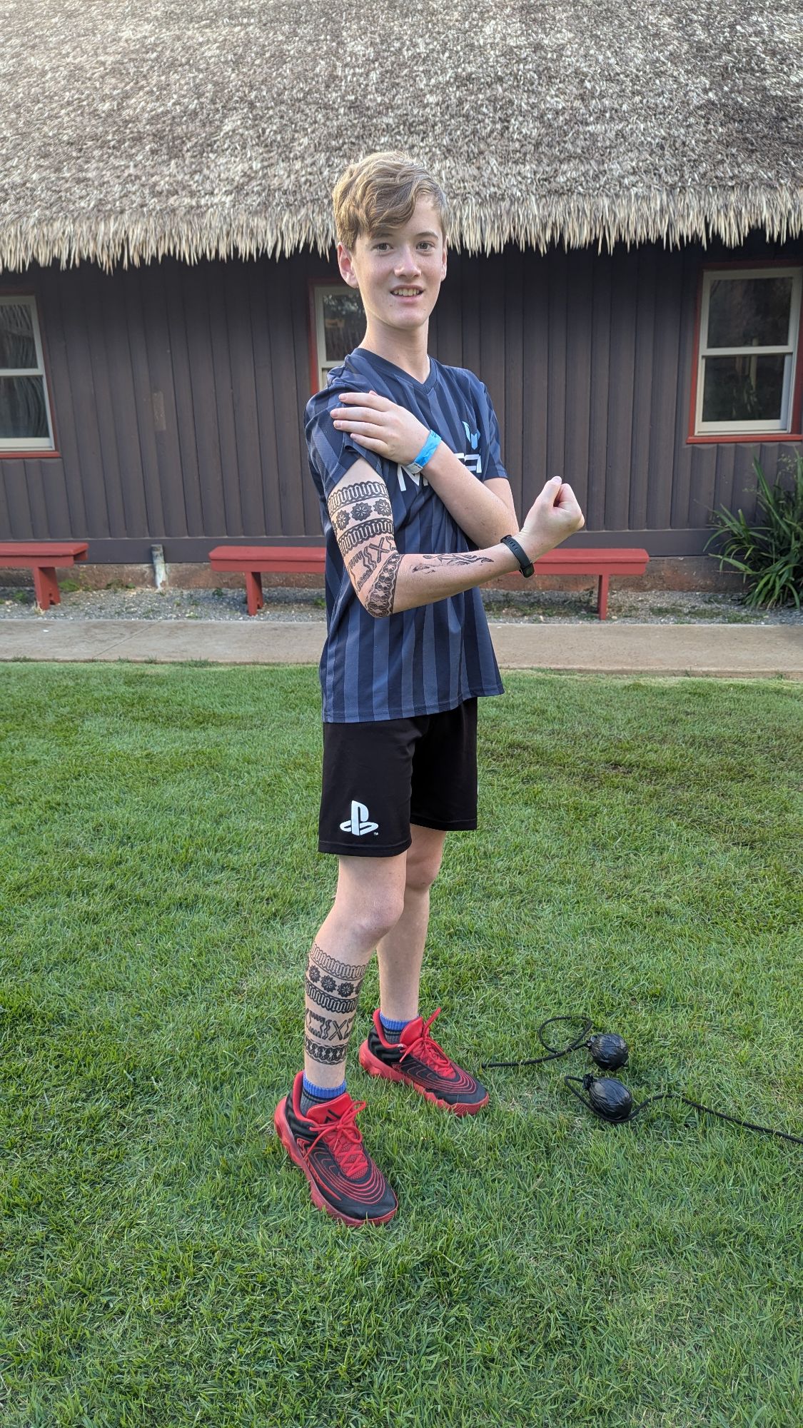 Teenager displaying temporary Maori-style tattoo on arm while standing on grass near traditional meeting house