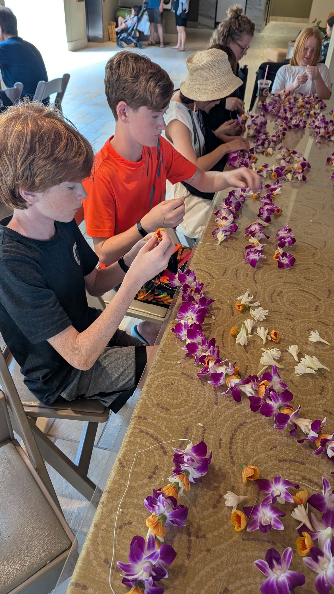 Children and adults stringing purple and white orchid leis at indoor activity station on cruise ship