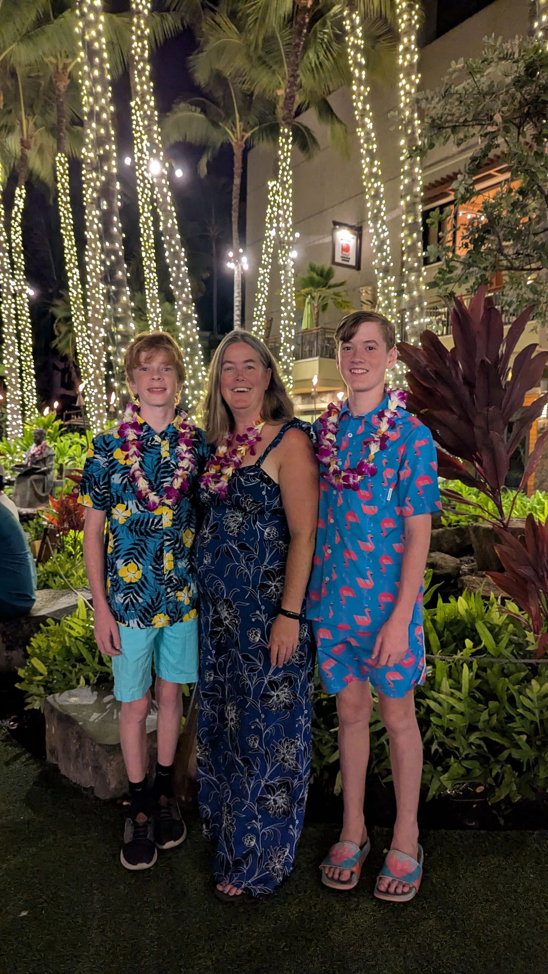 Marcy Bond with two teenage sons wearing colorful Hawaiian print shirts and lei garlands at night under illuminated palms