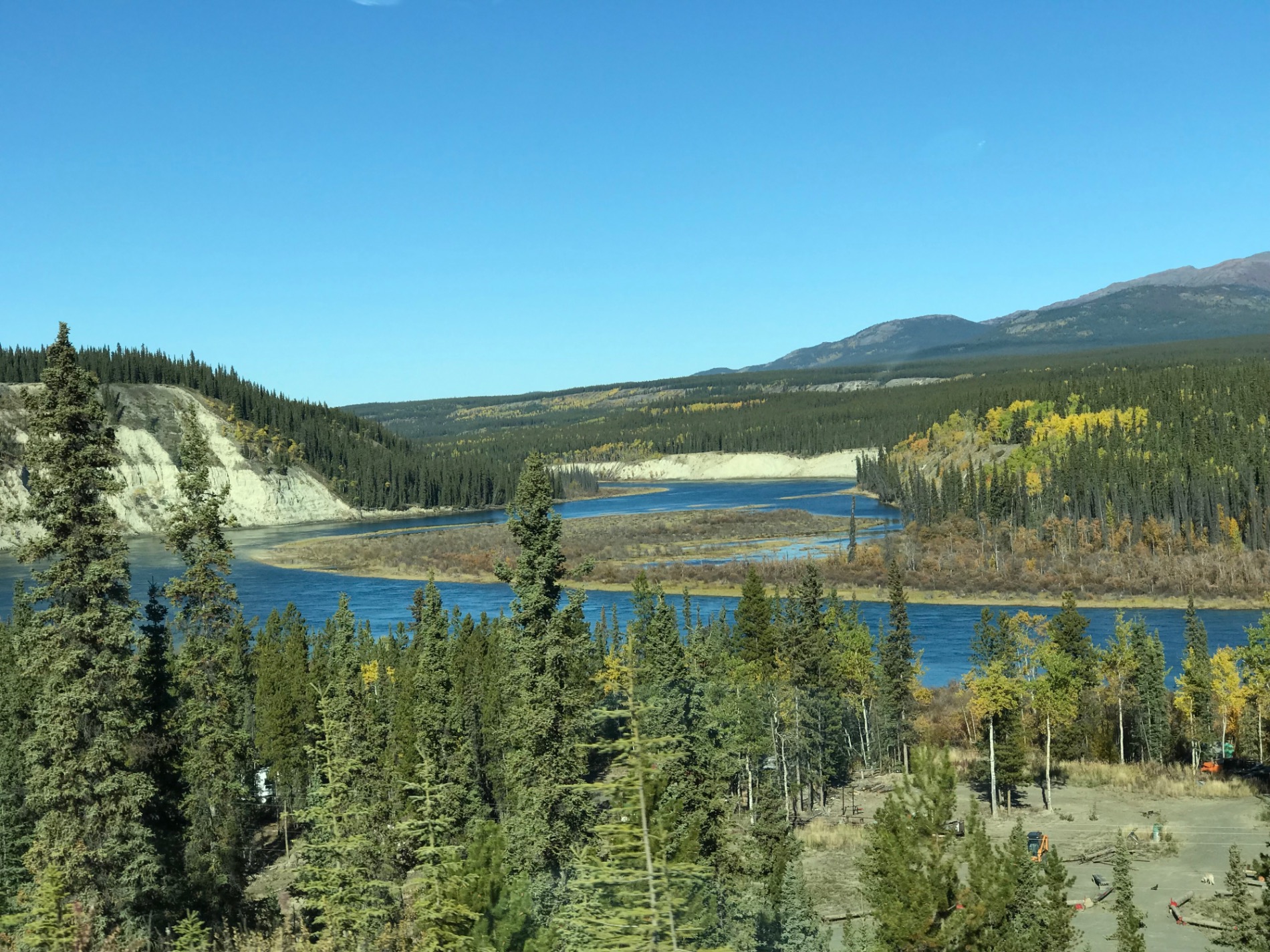 Mountain landscape with coniferous forest, blue river, and snow-capped peaks under clear sky in northern wilderness