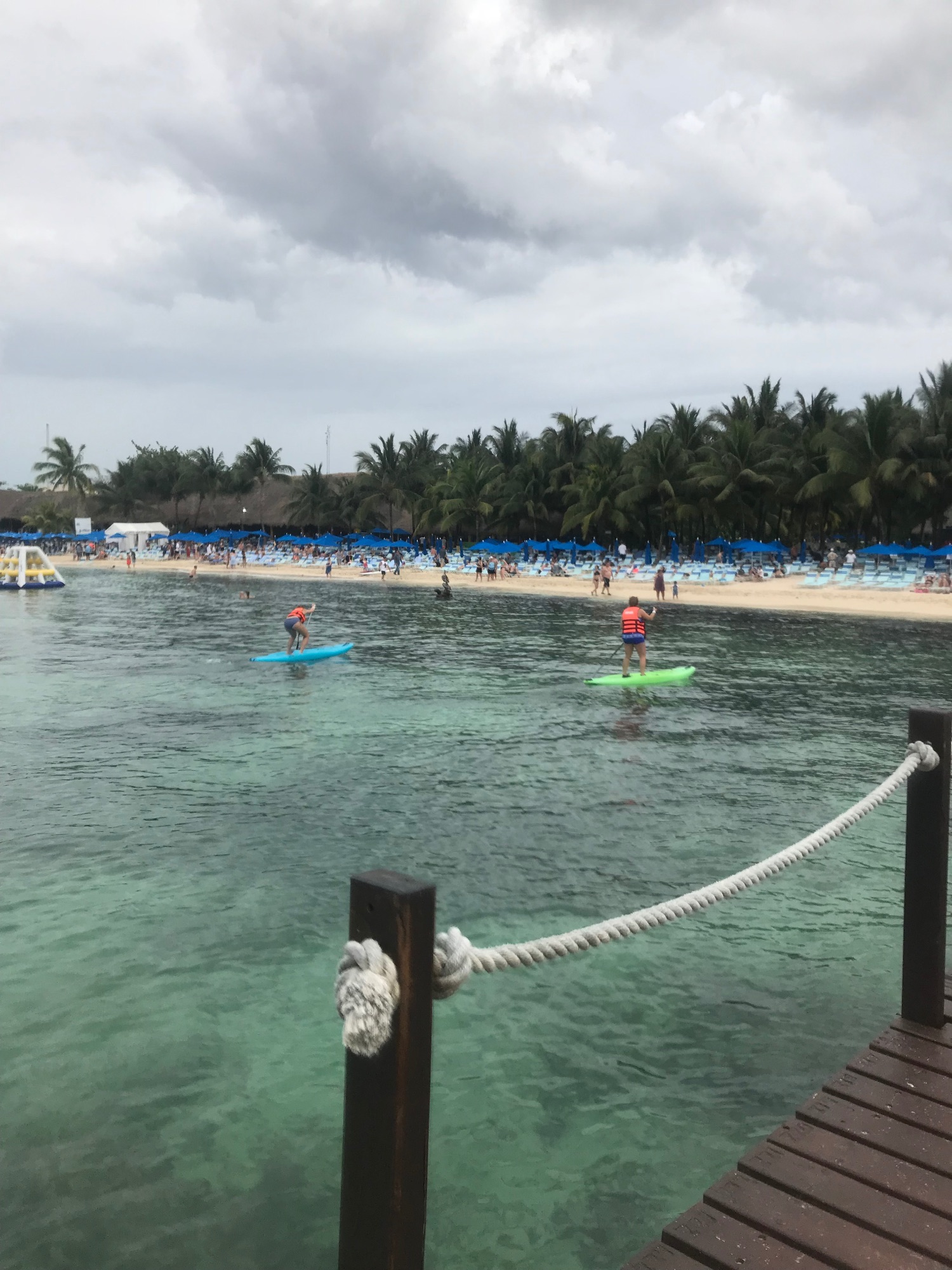 Beach scene with turquoise water, people paddleboarding and swimming, blue umbrellas on sand, and palm trees lining the shore