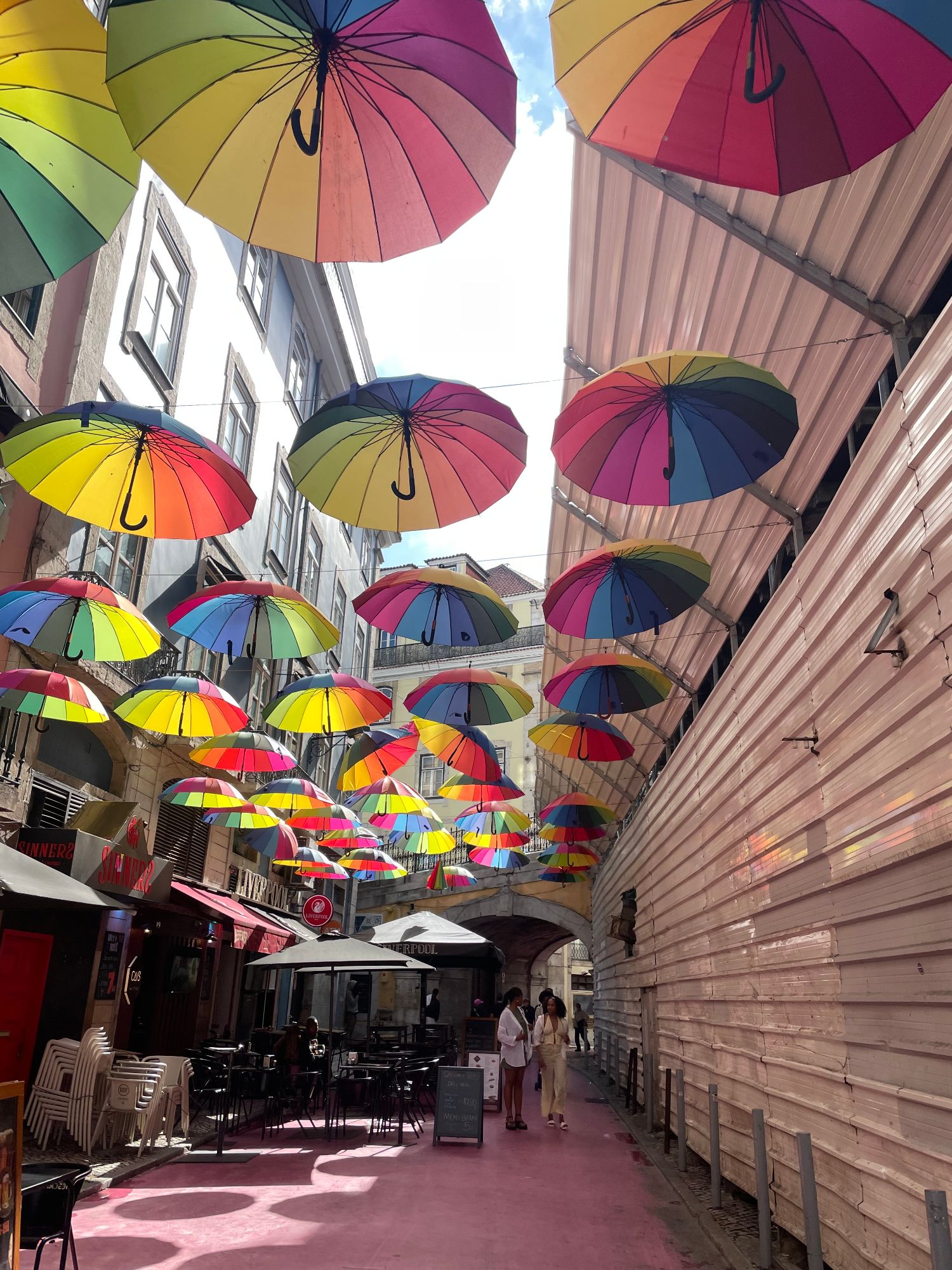 Colorful rainbow umbrellas suspended above a narrow European street with buildings and outdoor café seating
