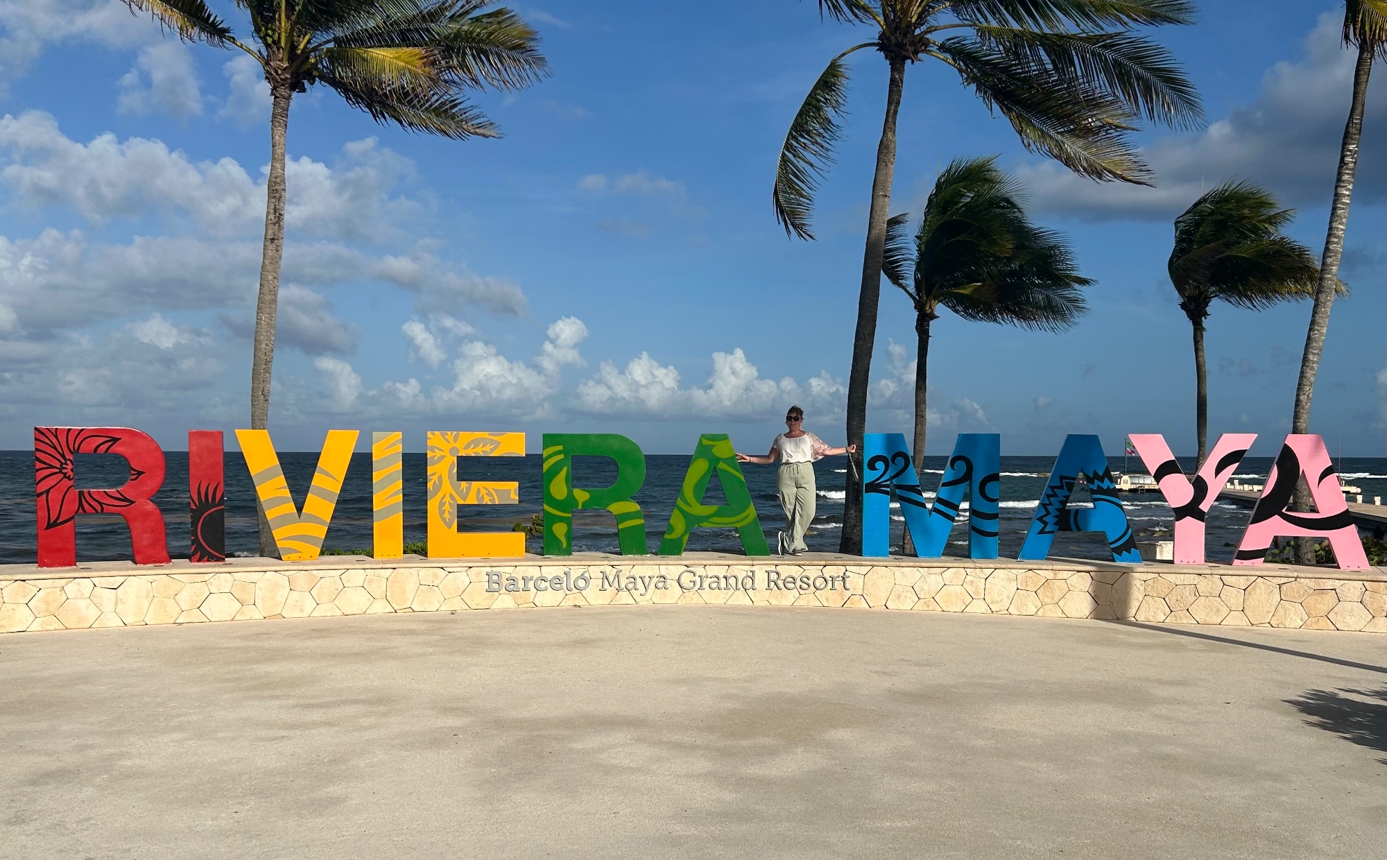 A woman poses at the colorful Riviera Maya sign at Barceló Maya Grand Resort with palm trees and ocean