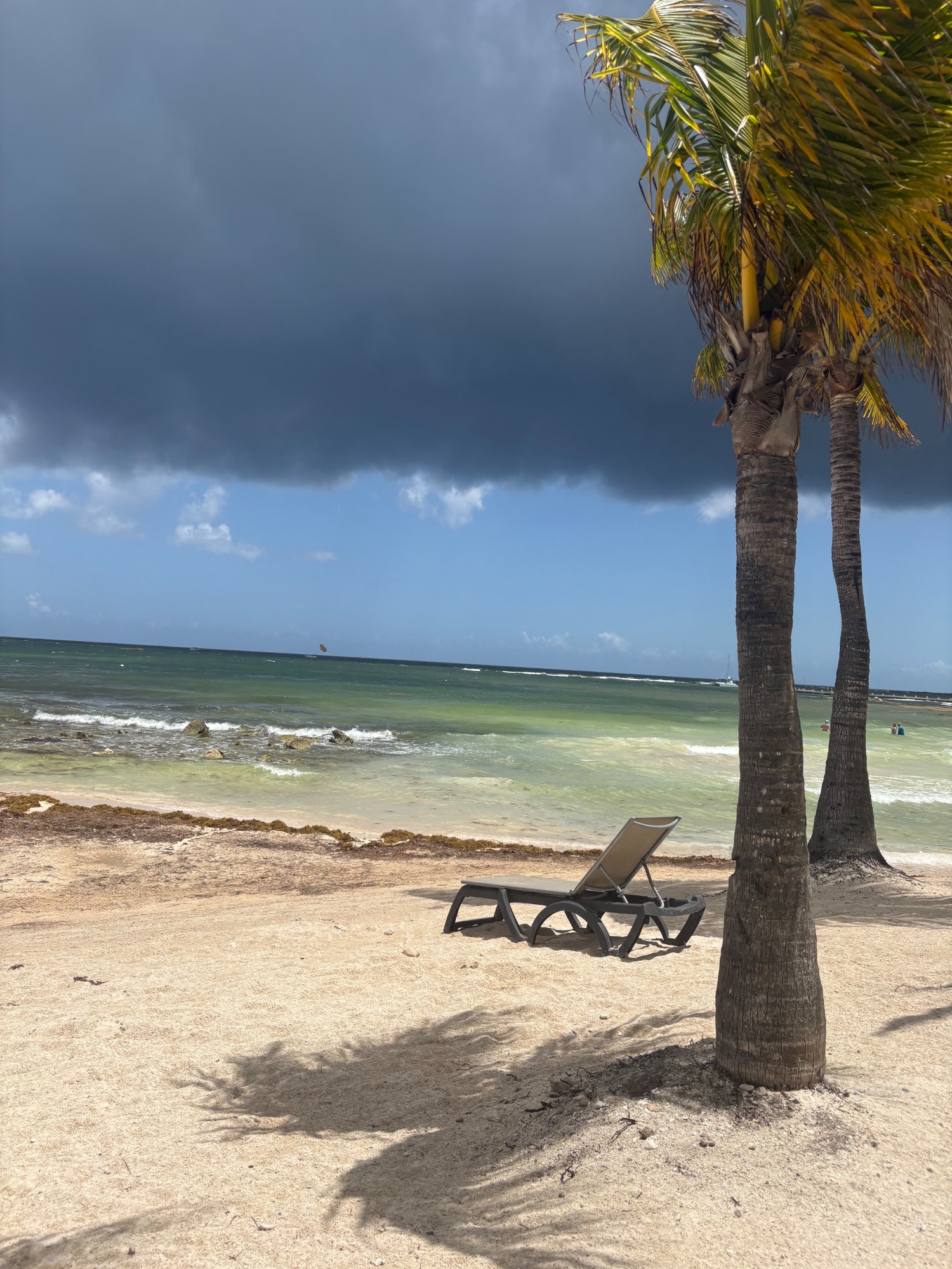 A lounge chair sits on sand beneath palm trees facing calm turquoise Caribbean water