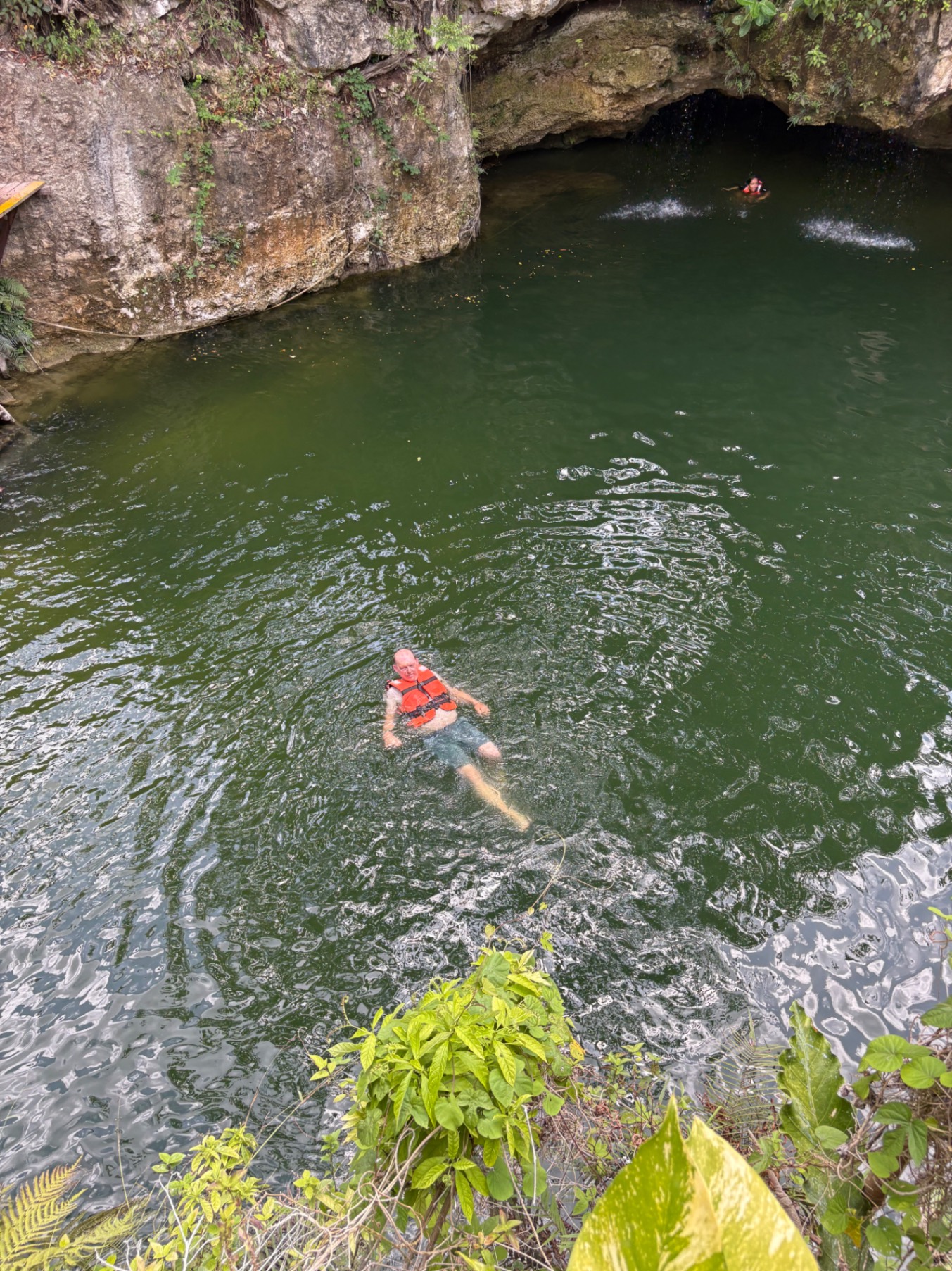 A man snorkels in clear green water inside a cenote cave with limestone formations overhead