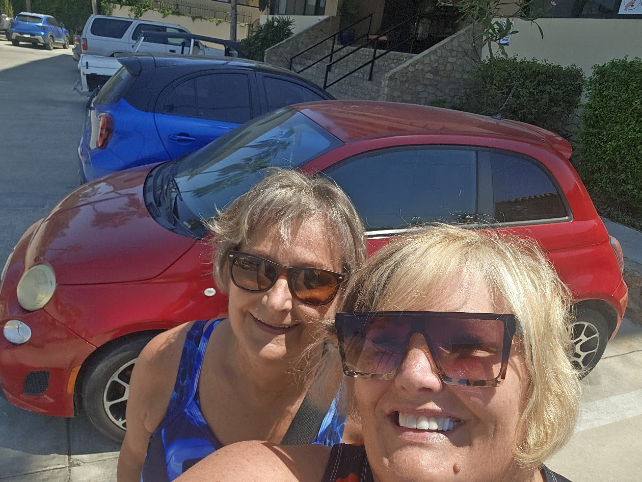 Two women posing together in front of red Fiat car on sunny street with blue car behind