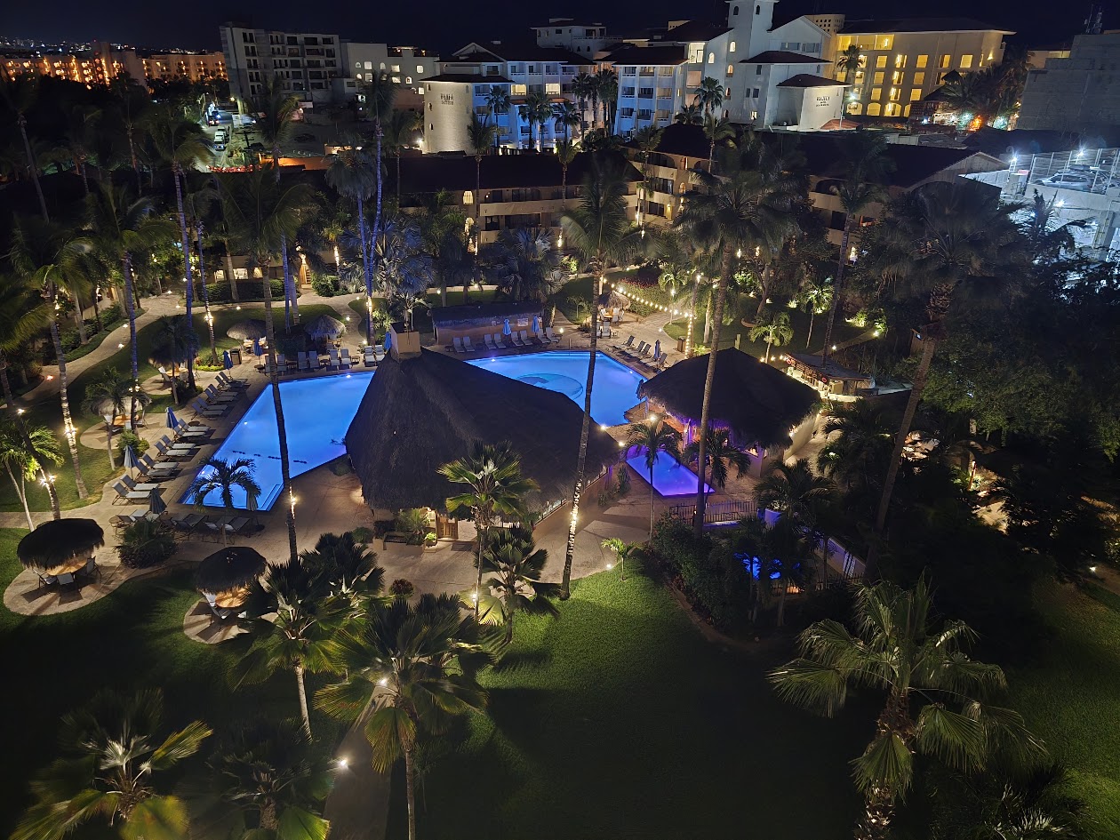 Illuminated resort pool complex at night with blue water, palm trees, and lit buildings in Cabo