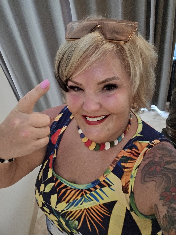 Joni Davis indoors wearing colorful patterned shirt and beaded necklace, pointing at camera