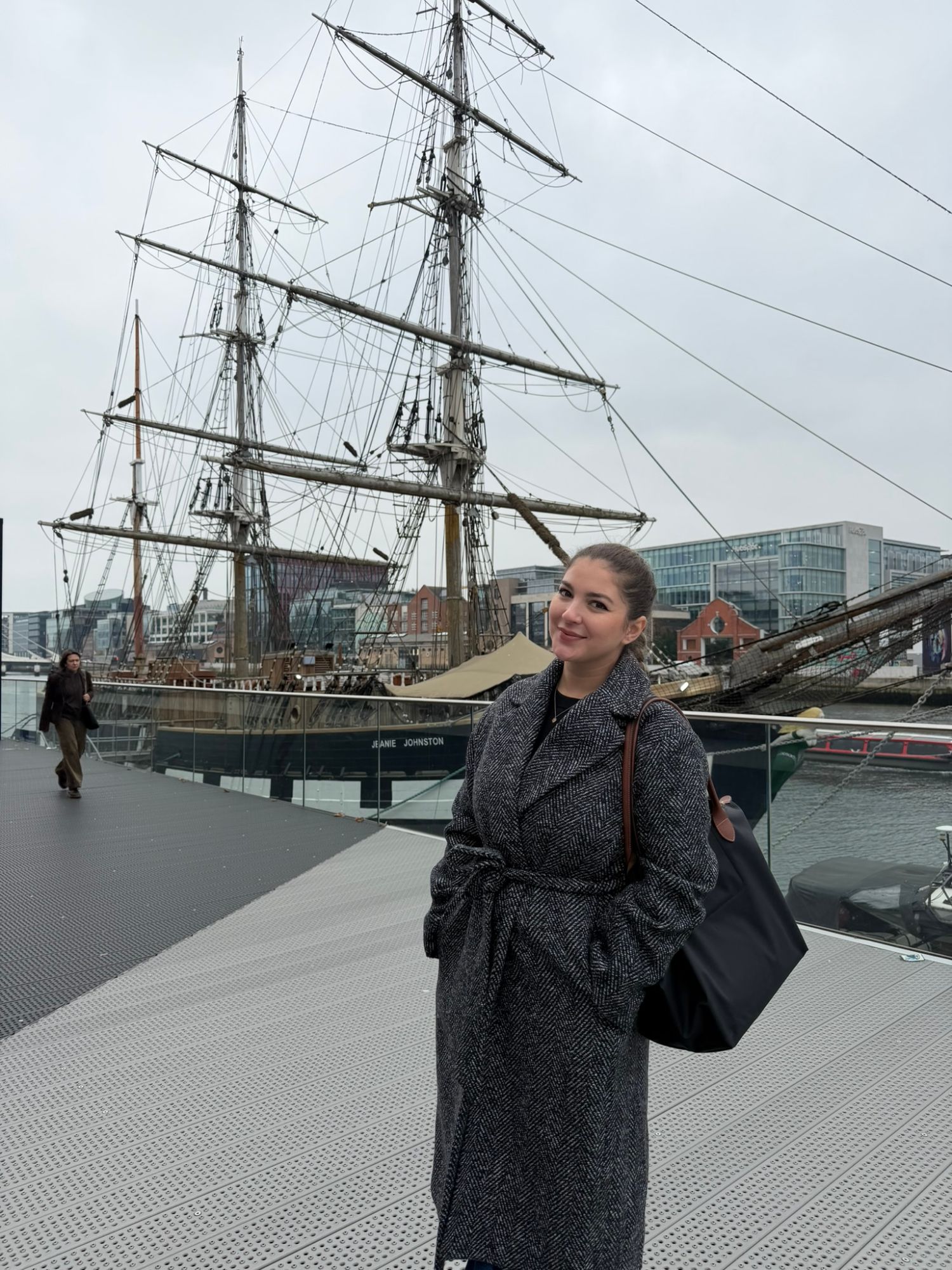 Woman in gray herringbone coat smiling at camera with tall ship Jeanie Johnston docked behind her