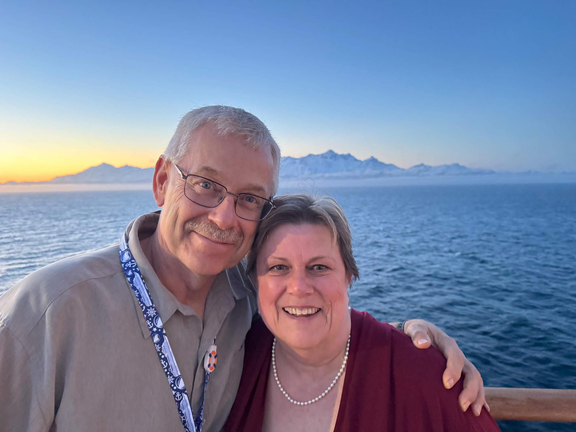Loreen Piehl-Wiedemann and a companion smile at sunset on a cruise ship deck with snow-capped mountains across the water