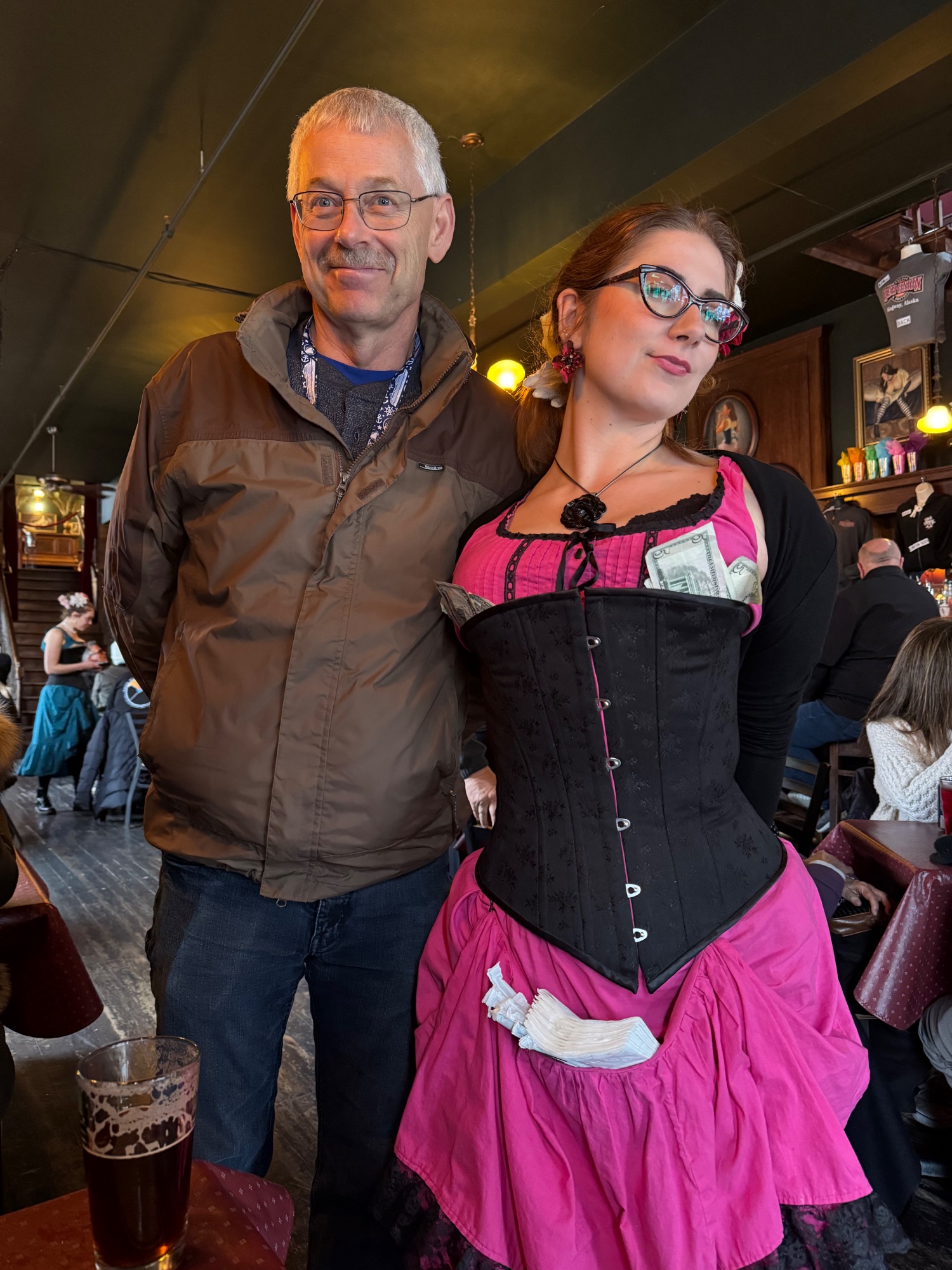 A man in glasses and brown jacket stands with a woman in a pink and black corset-style dress at an indoor event