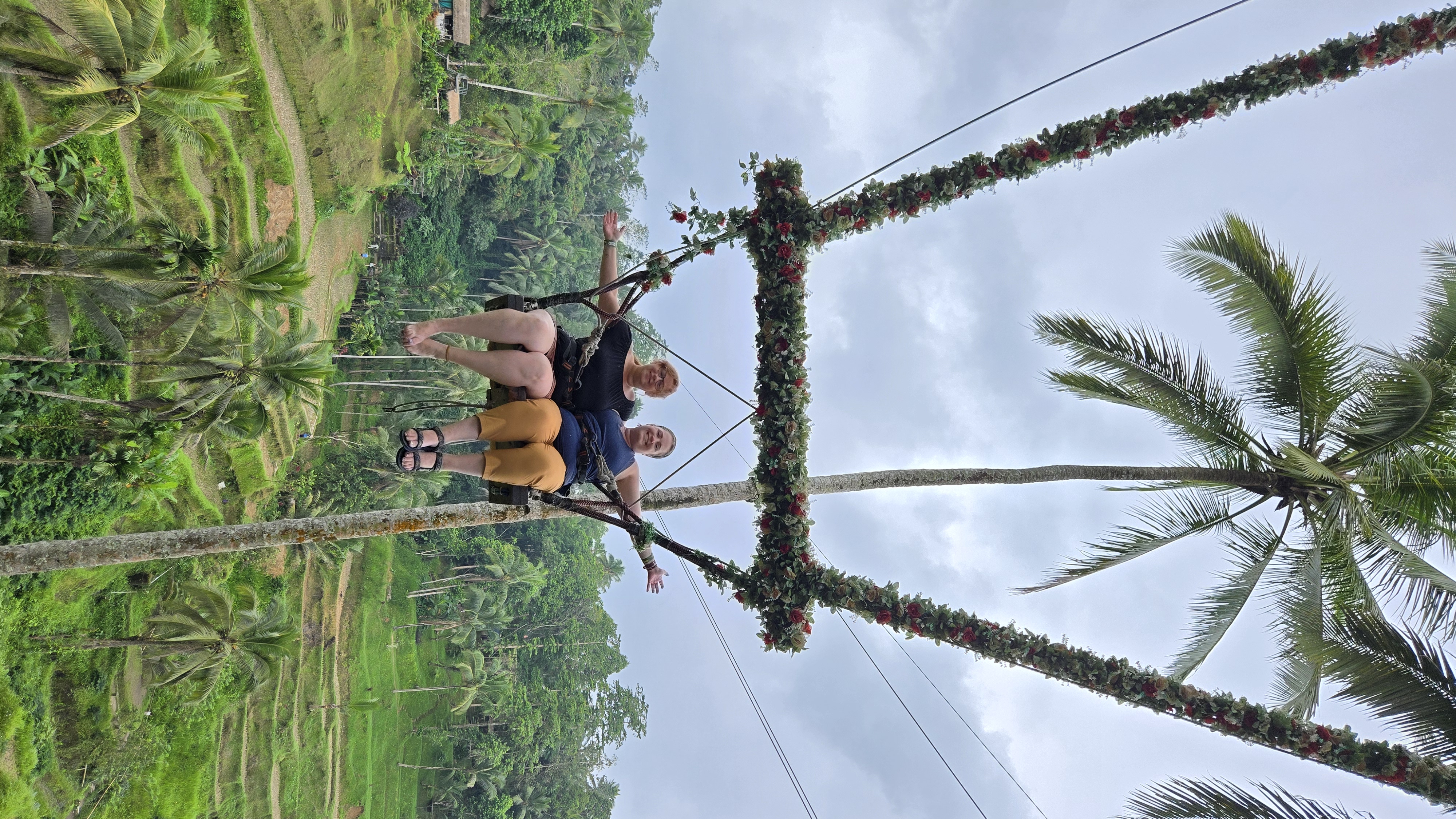 Two people sit on a swing suspended between two palm trees decorated with colorful flowers, overlooking green agricultural fields