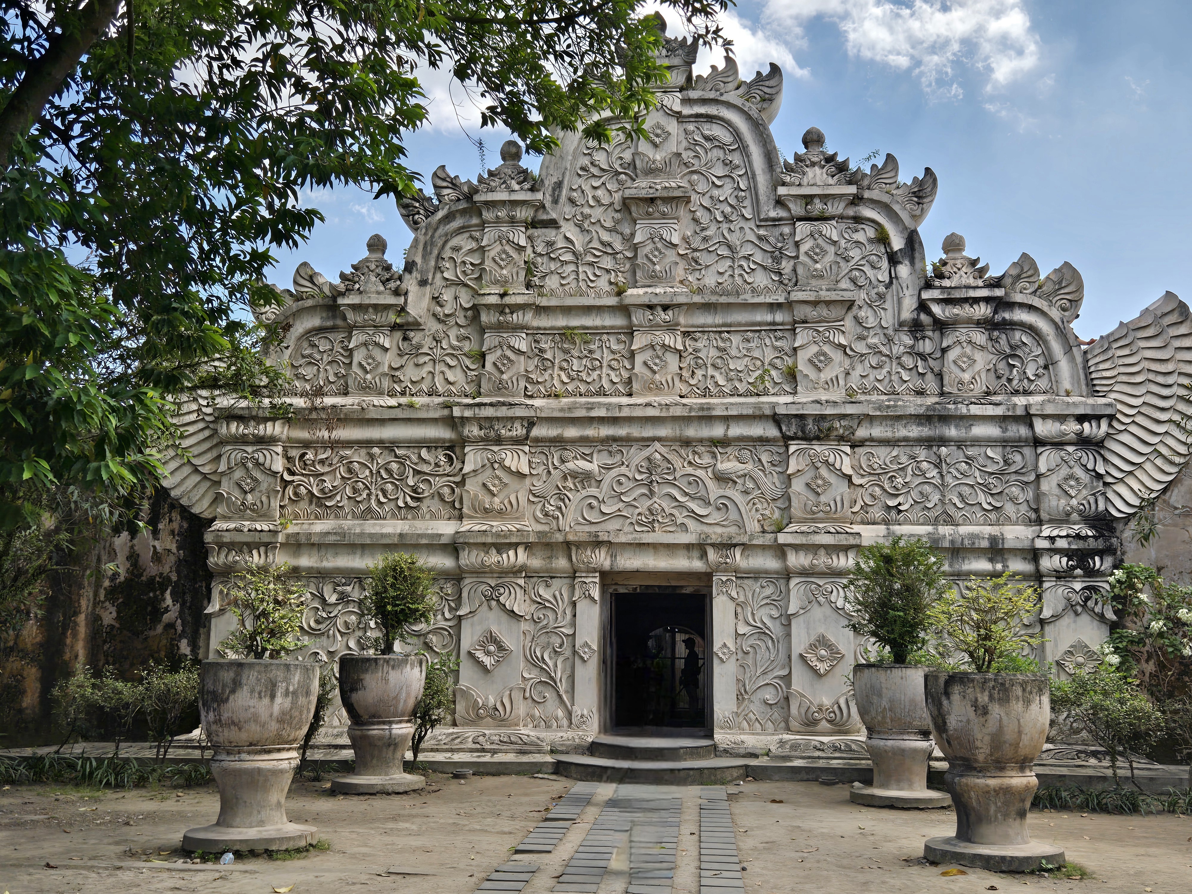 An ornately carved stone temple structure with intricate relief work and a central doorway, flanked by decorative urns