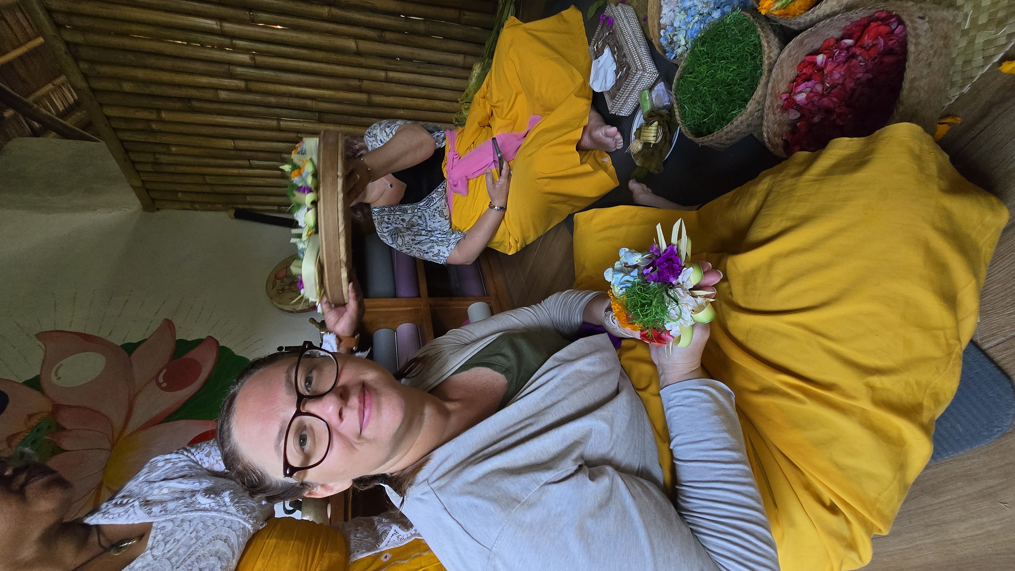 Kelly holds a colorful flower arrangement at a workshop, wearing yellow fabric and surrounded by baskets of vibrant flowers and greenery