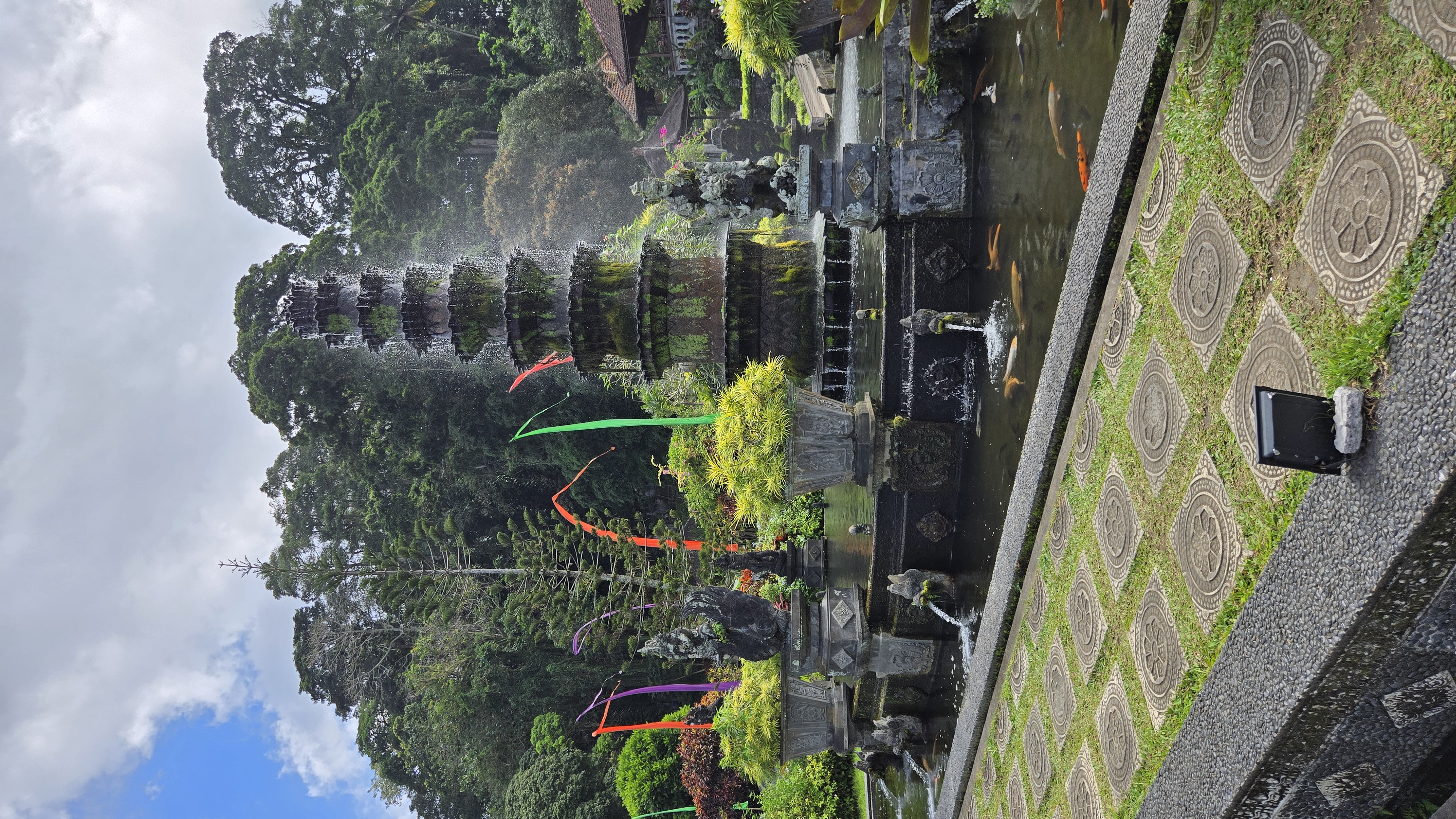 A stone temple tower with multiple tiers covered in moss, surrounded by lush forest with colorful ribbons hanging from nearby structures