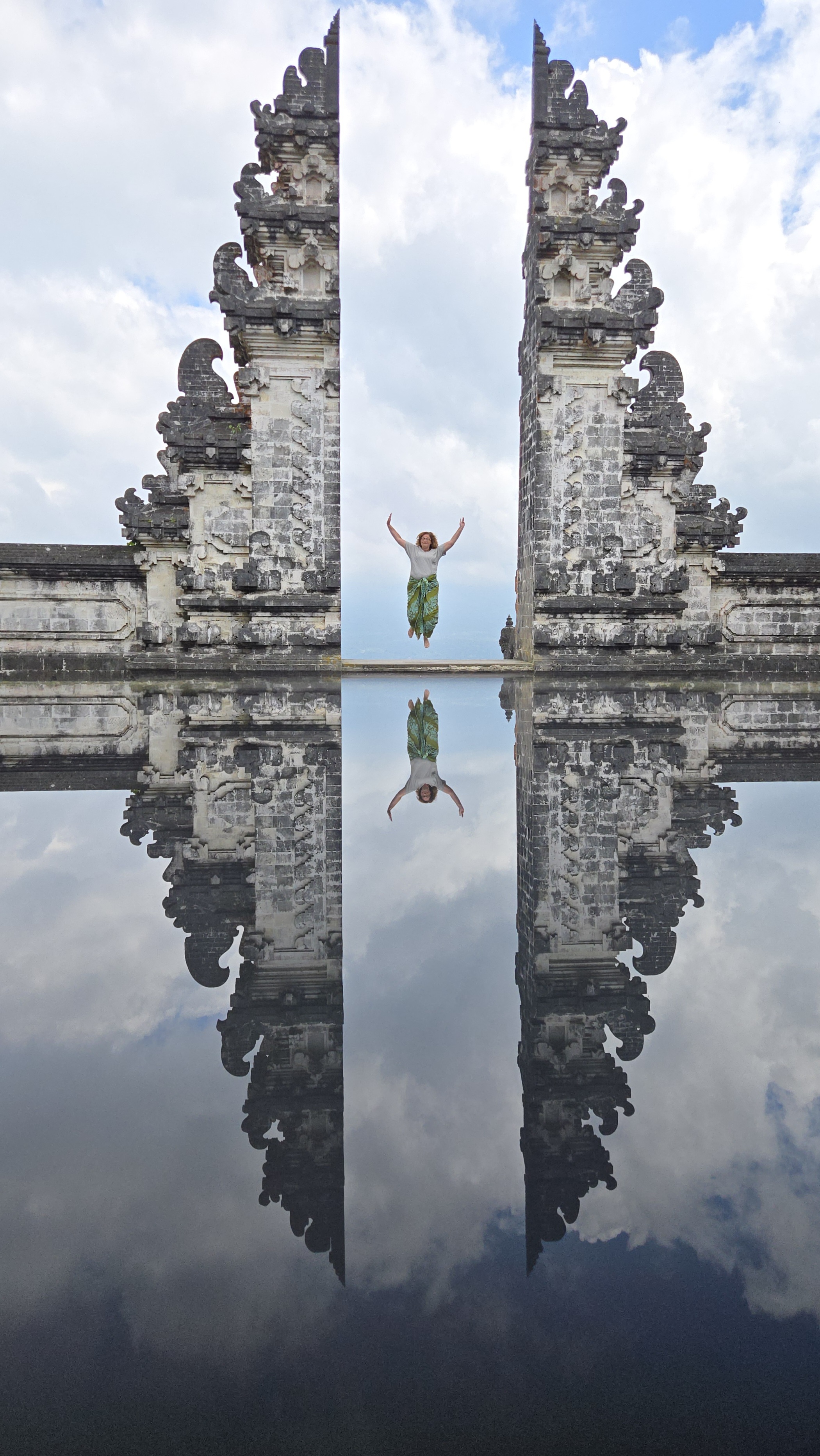 A woman jumps with arms raised between two ornate stone temple gates reflected in still water below